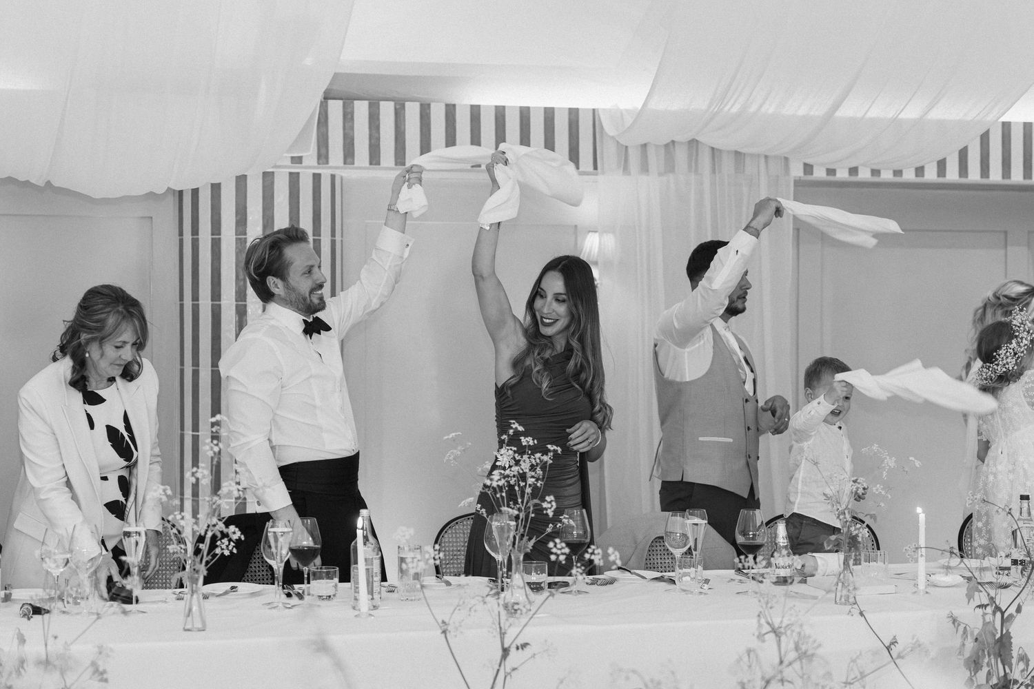 Wedding party waves white napkins in celebration at elegantly decorated reception table in marquee.