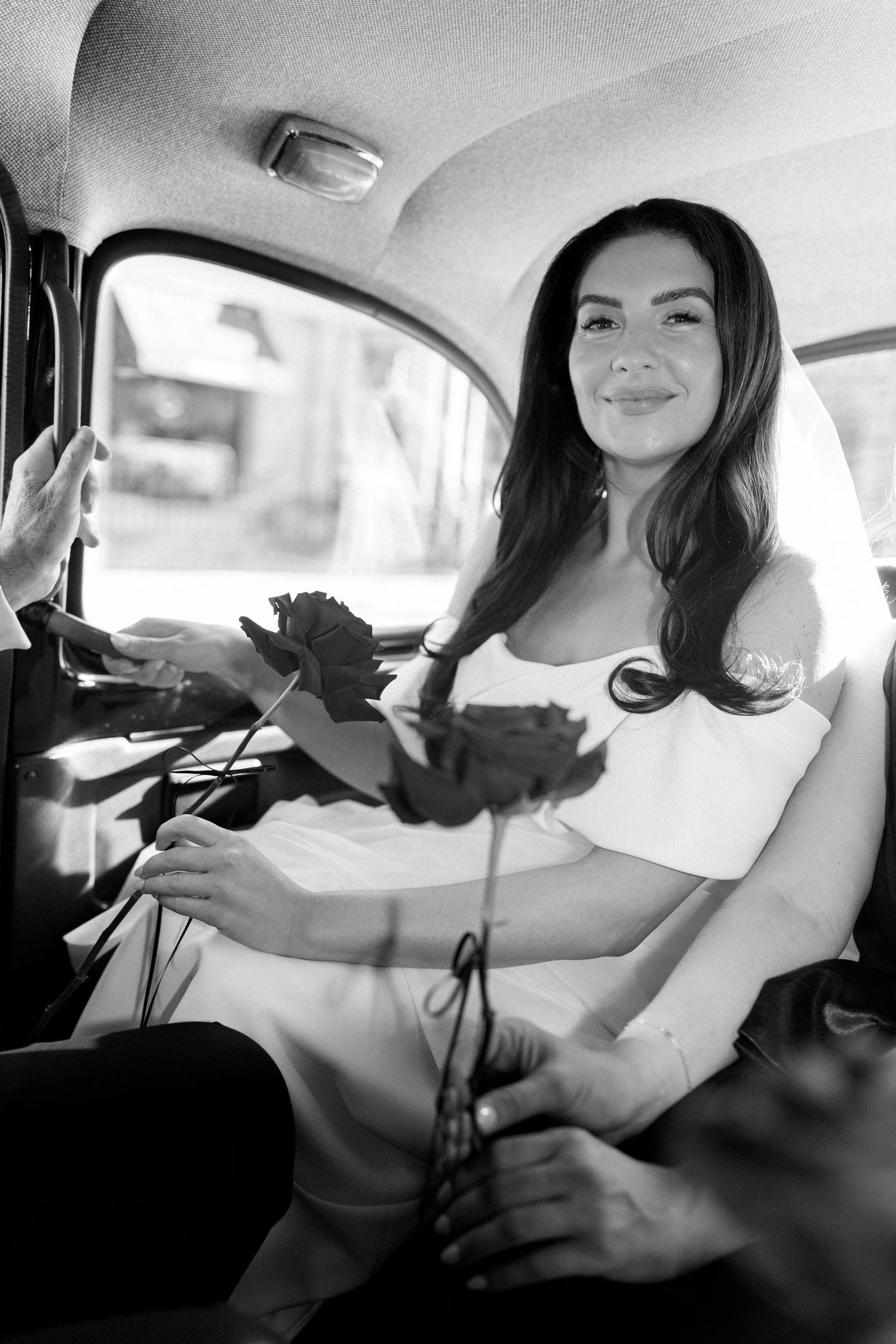 Smiling bride in an off-shoulder white dress sits in the back of a vintage taxi holding a red rose in this black and white wedding photo.