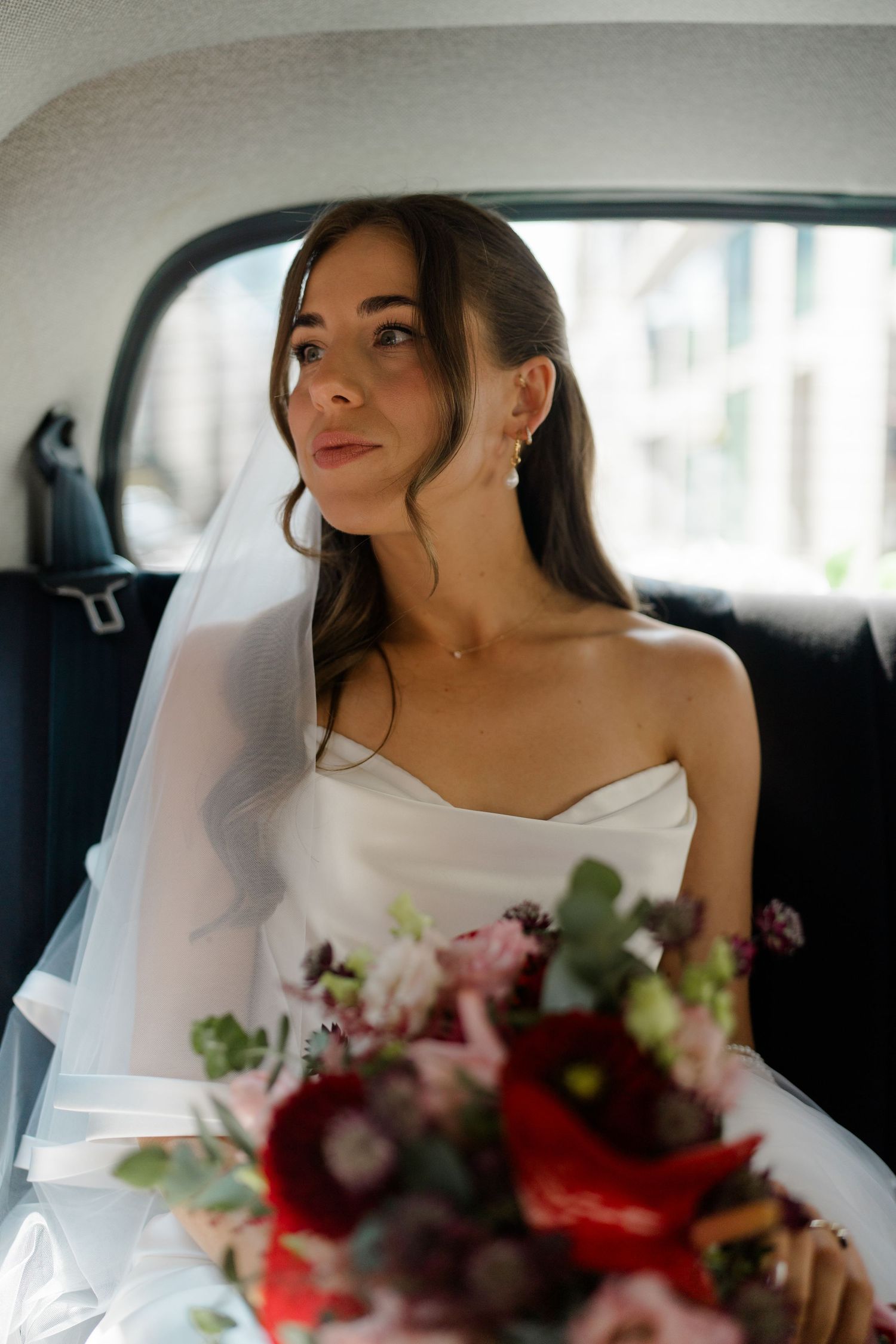 Bride in strapless white gown holding red bouquet sits in car backseat wearing veil and pearl earrings.