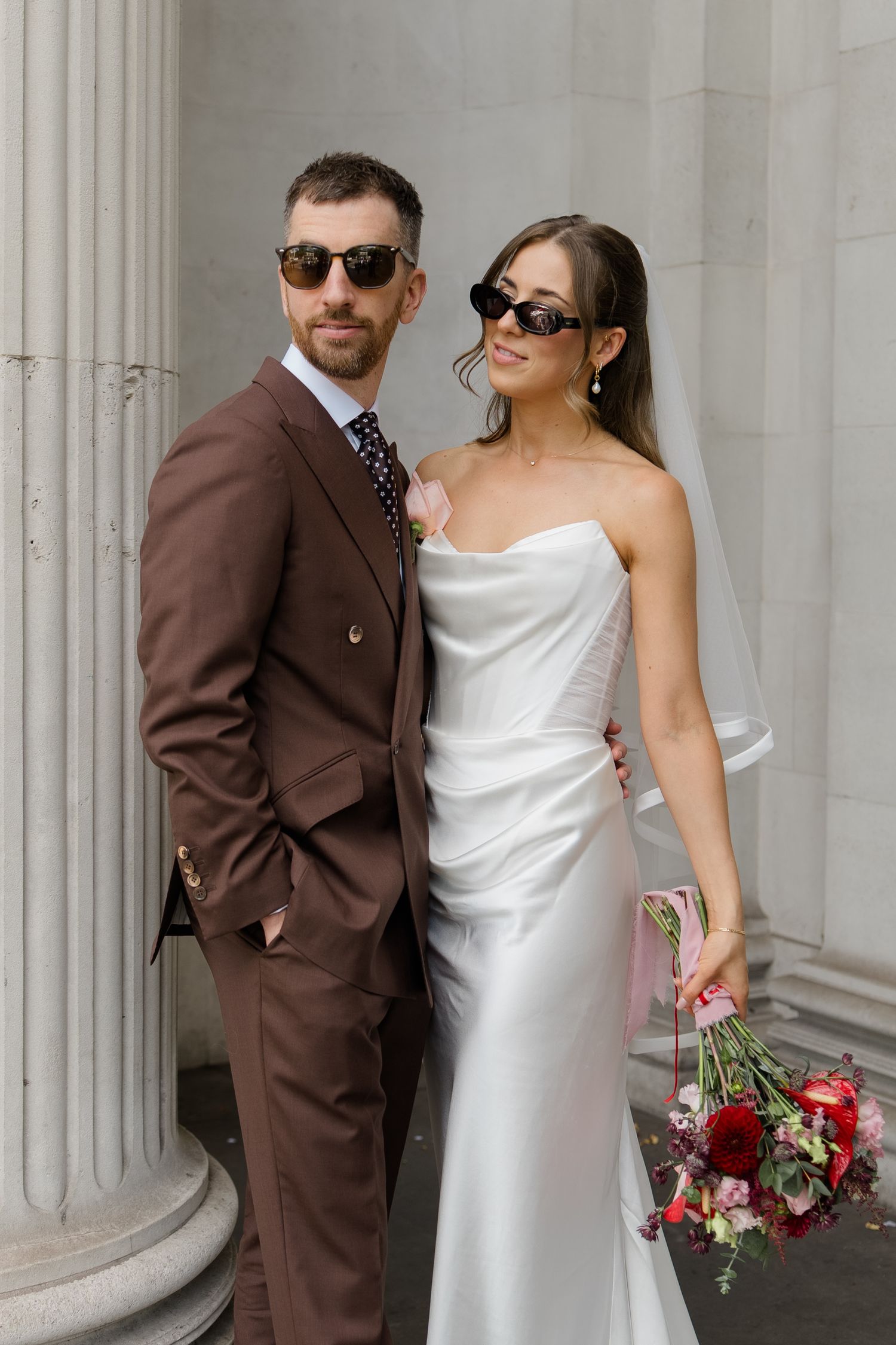 Stylish bride and groom posing in sunglasses, she in white satin gown, he in brown three-piece suit.