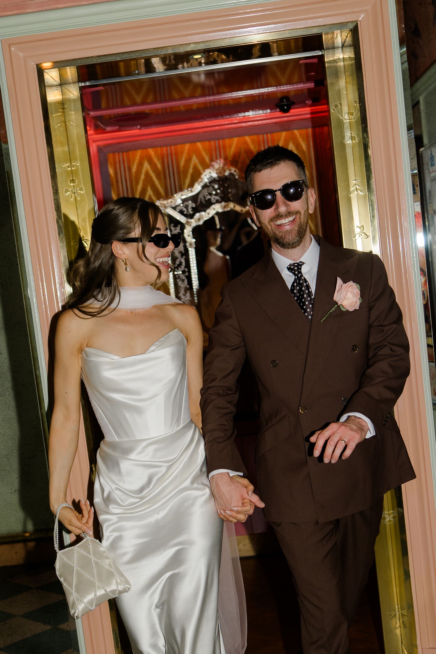 Stylish bride and groom in sunglasses holding hands exiting a glamorous venue entrance.