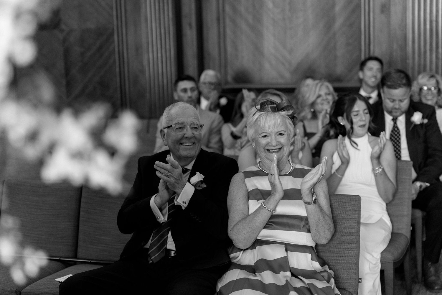 Black and white photo of joyful wedding guests applauding during ceremony in elegant wood-paneled room.