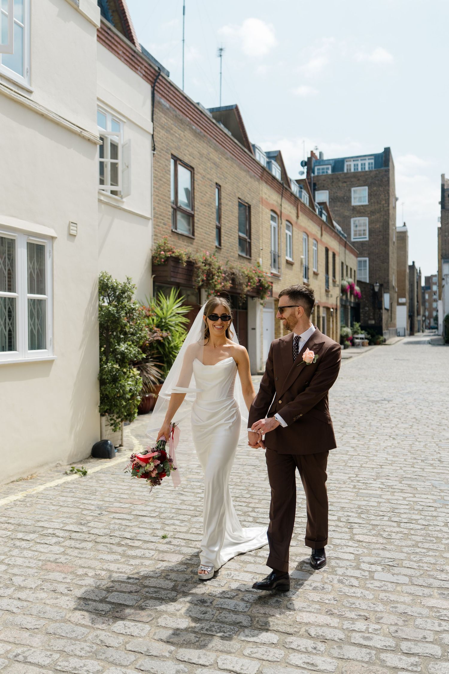 Bride in white gown and groom in brown suit walk cobblestone mews street on sunny wedding day.