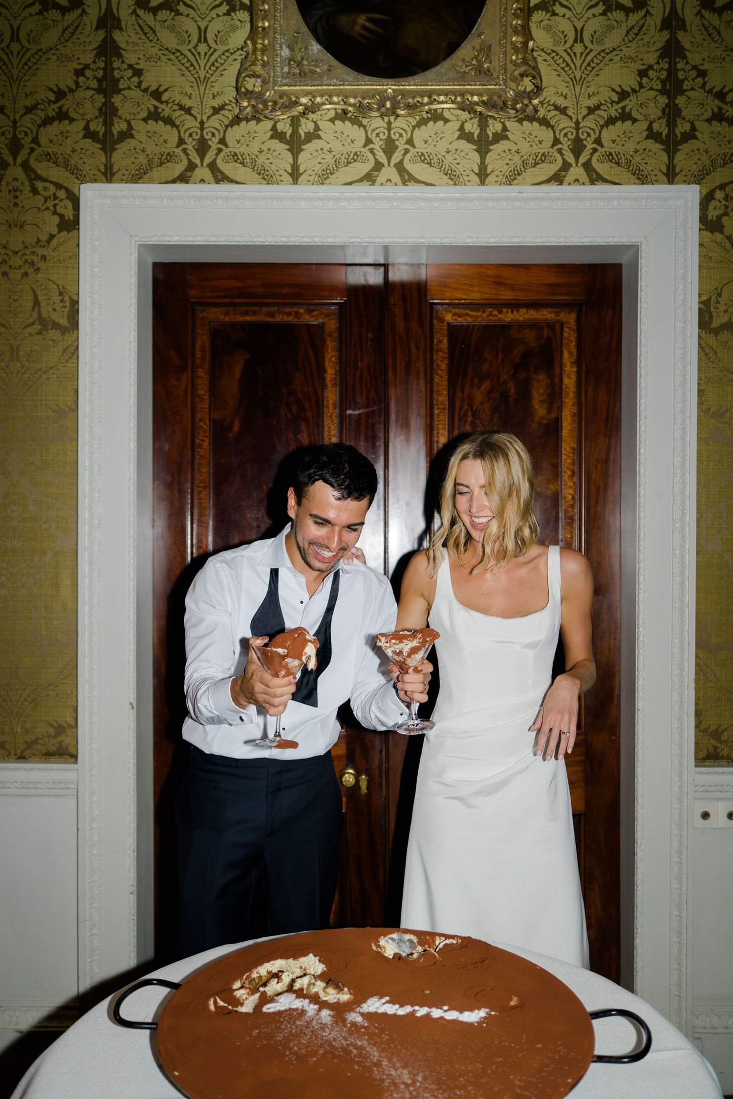 Bride and groom laughing while cutting wedding cake in ornate gold wallpapered room.
