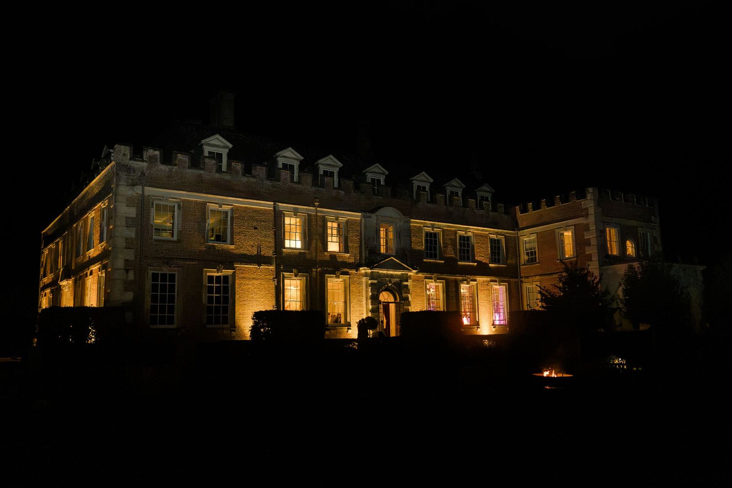 A grand historic English manor house dramatically illuminated with warm golden lights at night.