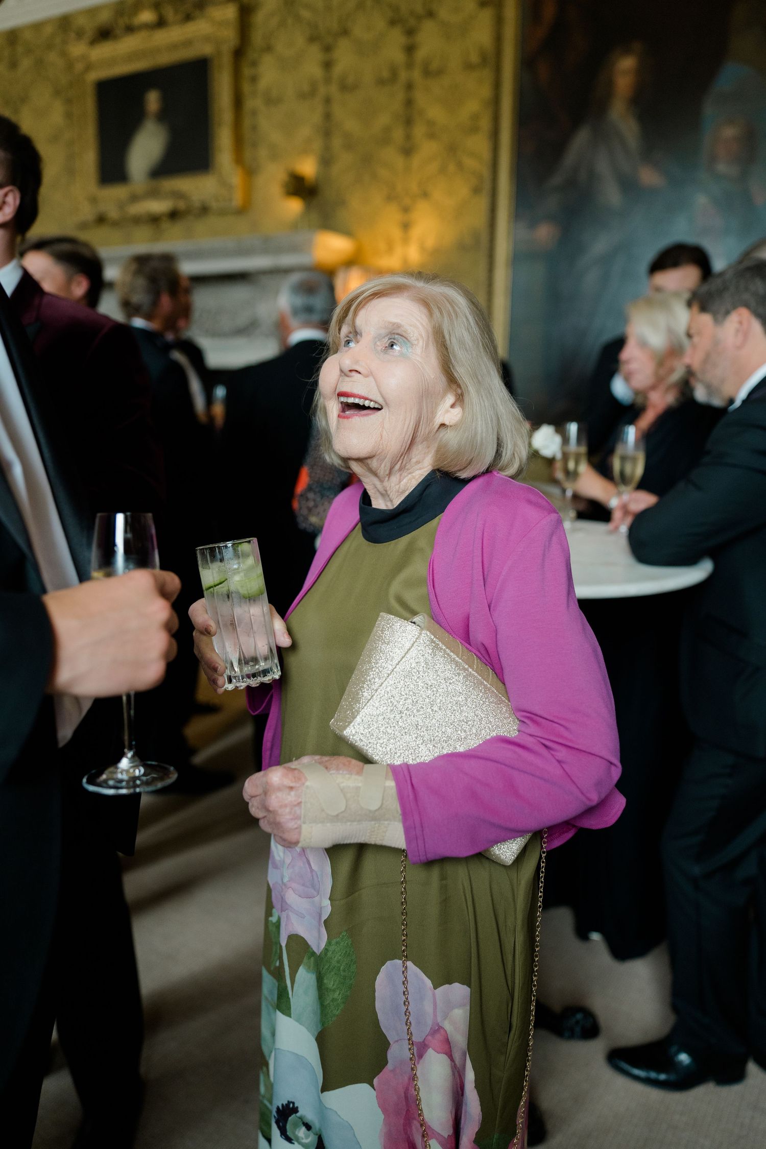 Elderly woman in pink cardigan laughing at elegant party, holding drink and clutch purse.