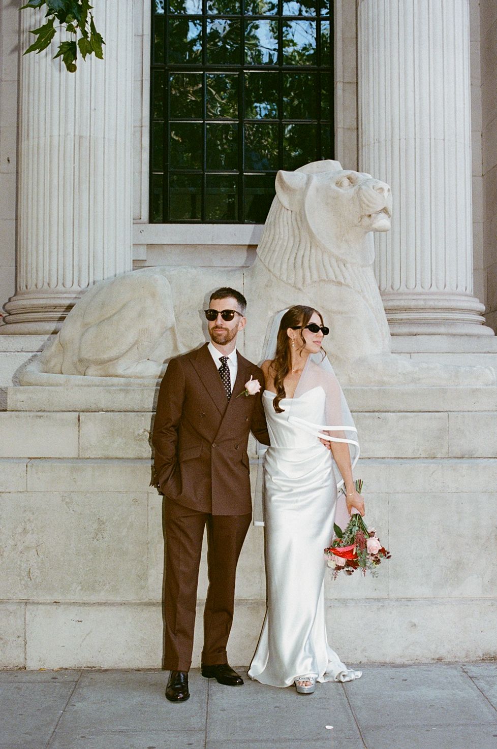 Bride and groom in sunglasses pose before stone lion sculpture and classical columns.