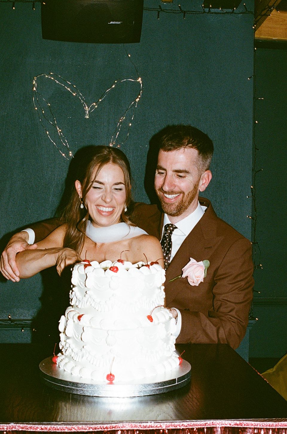 Bride and groom cutting white tiered wedding cake with sparkler heart backdrop.
