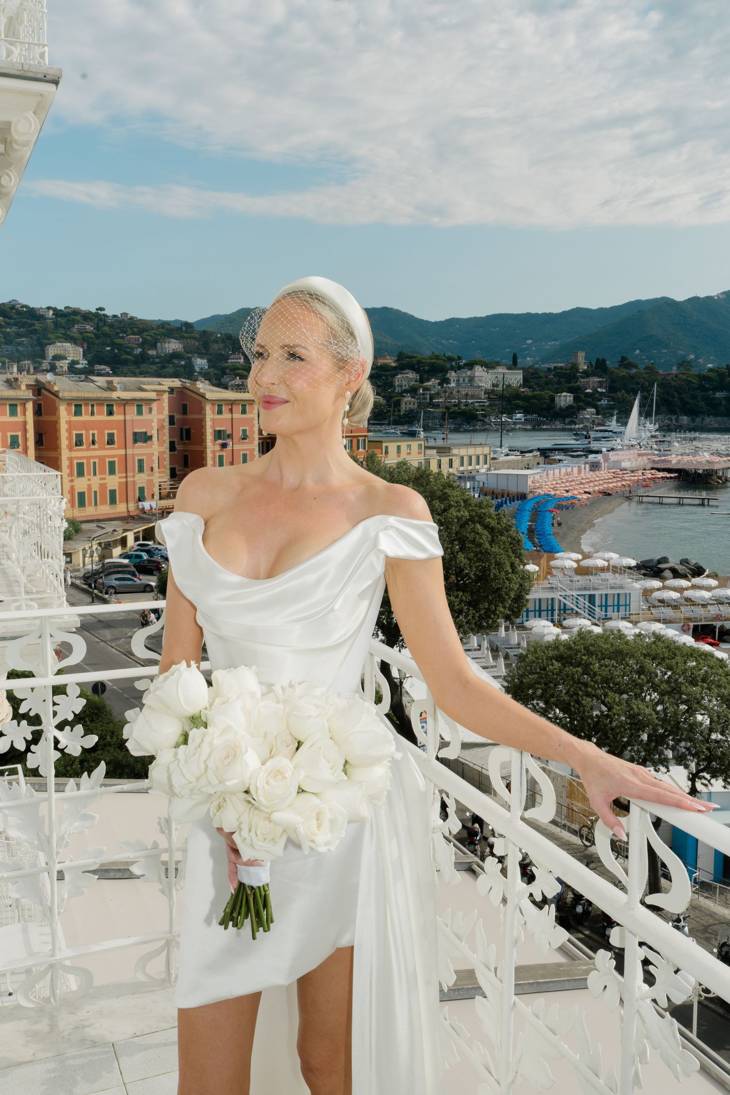 Elegant bride in off-shoulder white mini dress holding white peony bouquet on ornate balcony in Italian coastal town.