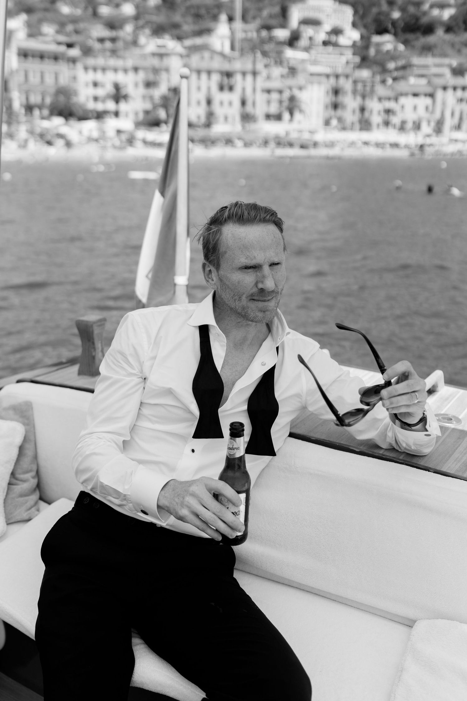 Stylish man in white tuxedo shirt holding beer bottle on yacht near Mediterranean coastal town.