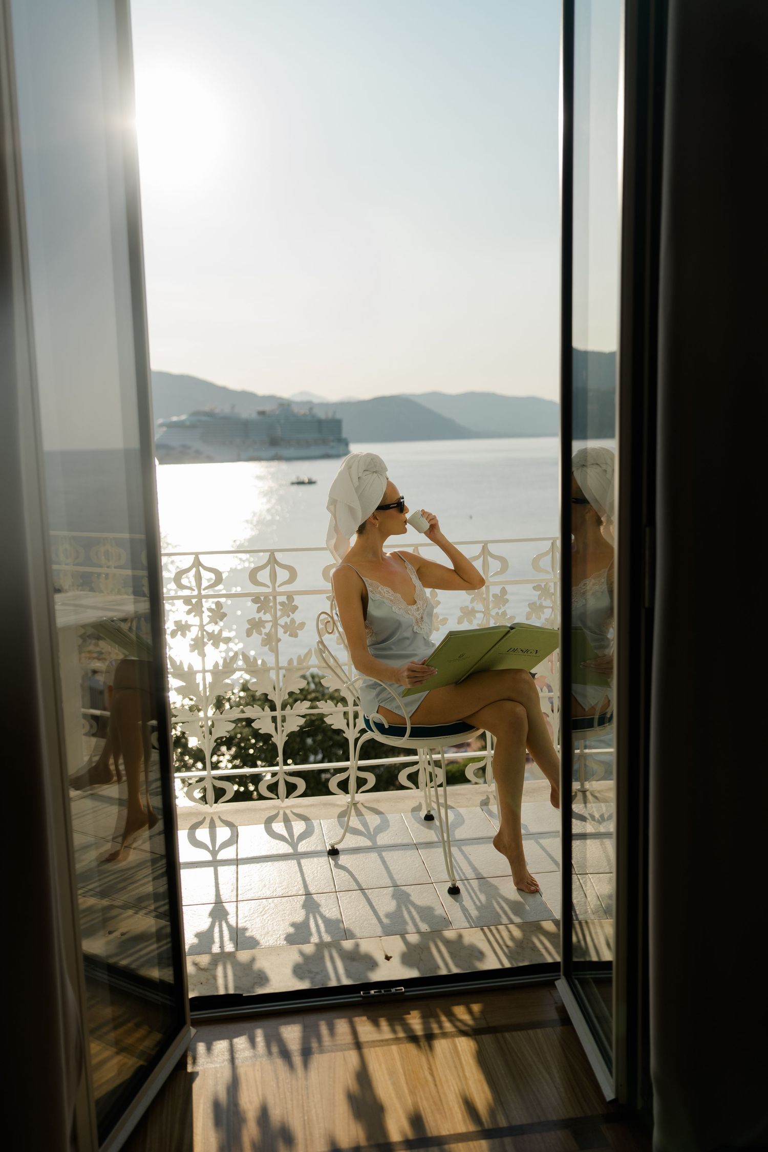 Woman with towel turban drinks coffee while reading on elegant white balcony overlooking calm sea.