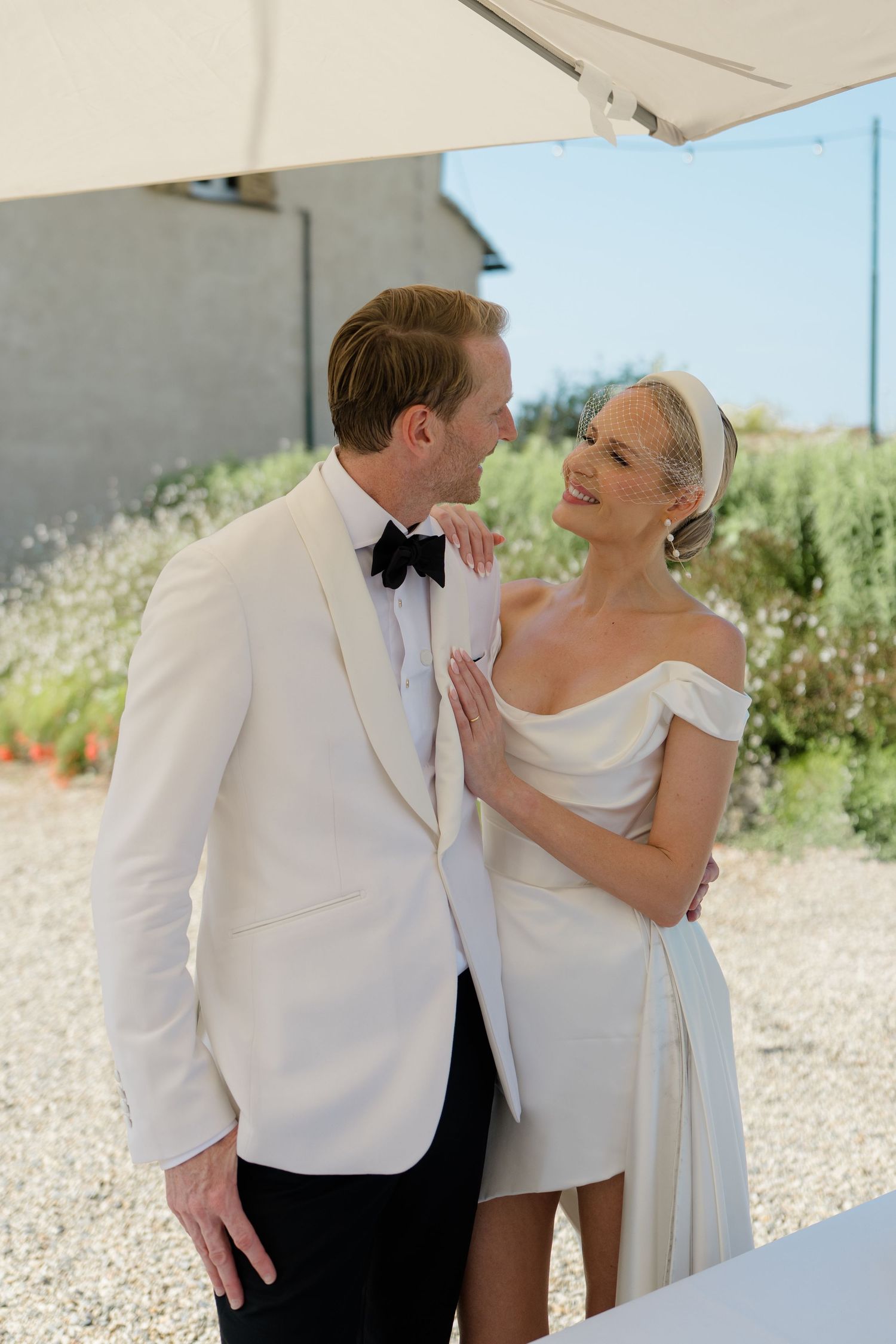 Bride and groom in elegant white wedding attire smiling at each other outdoors in sunny setting.