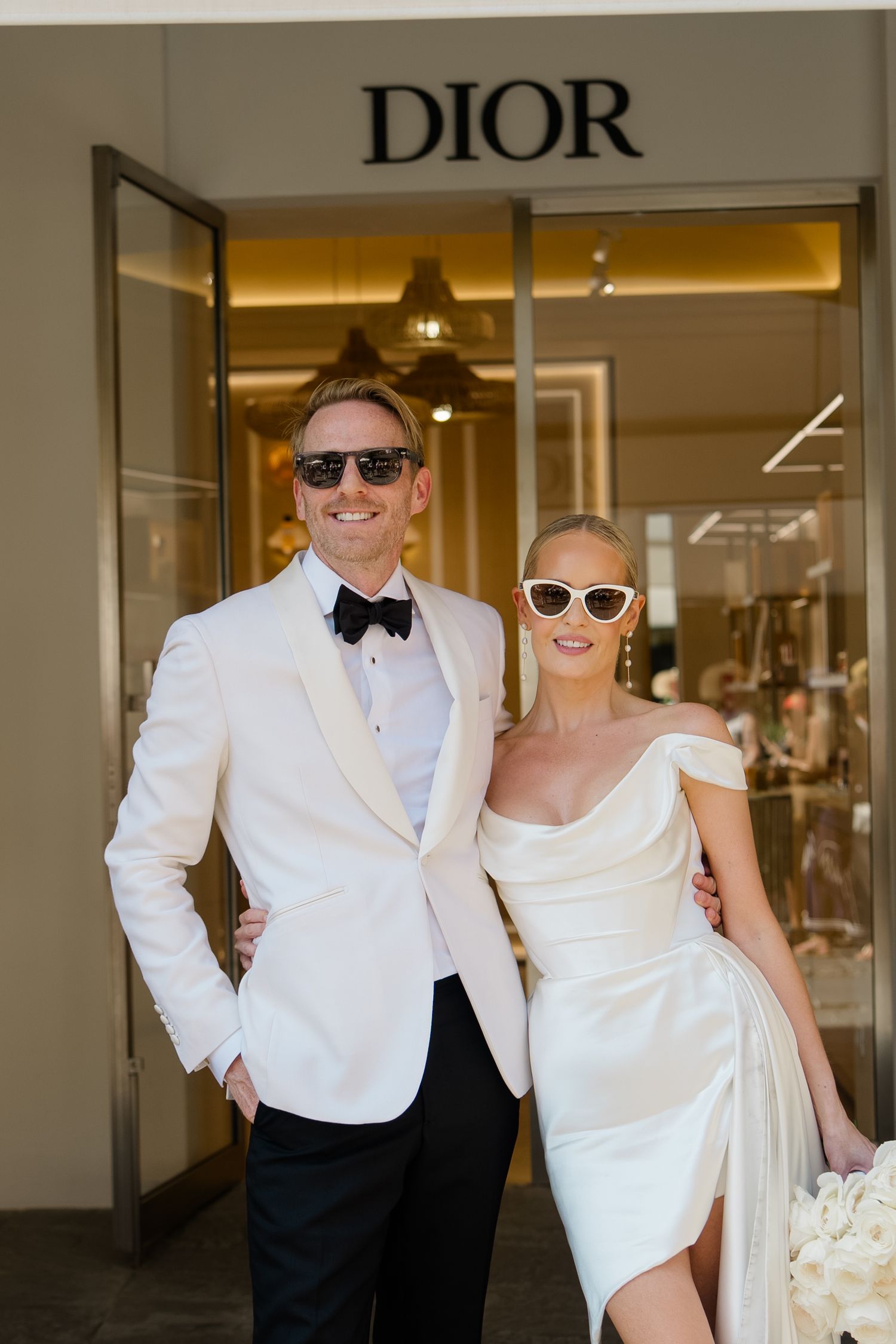 Chic bride and groom in matching white outfits and sunglasses pose smiling outside Dior boutique.