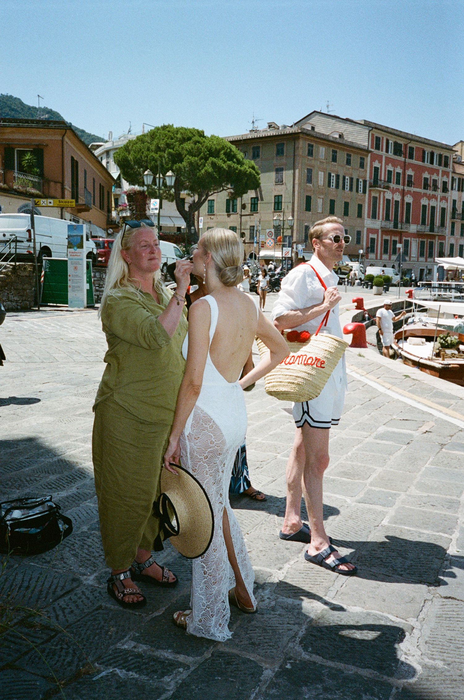 Three tourists stroll through sunny Italian harbor town, woman in white lace dress carrying straw hat.