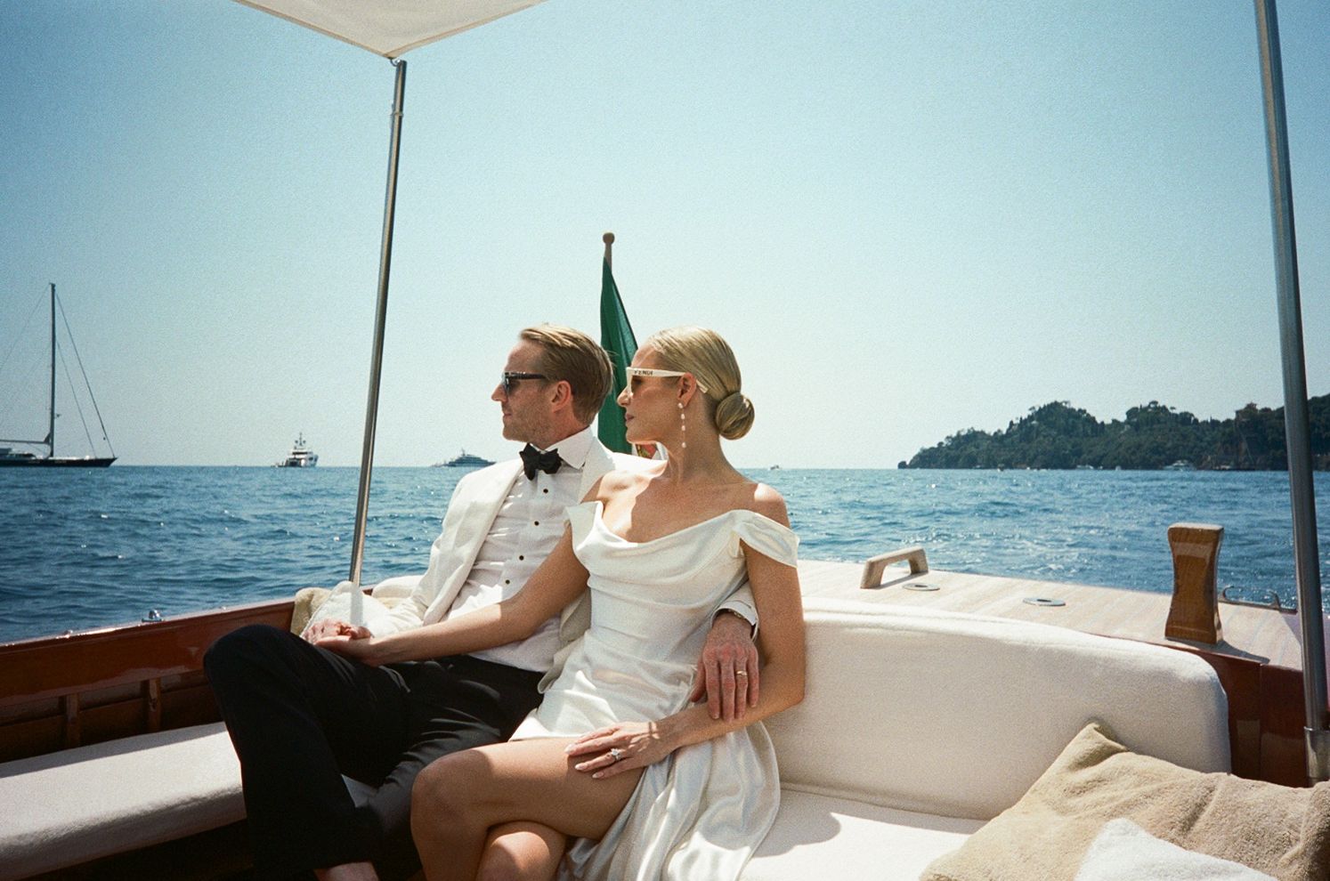 Elegant bride and groom in formal attire seated on luxury wooden boat on blue Mediterranean sea.