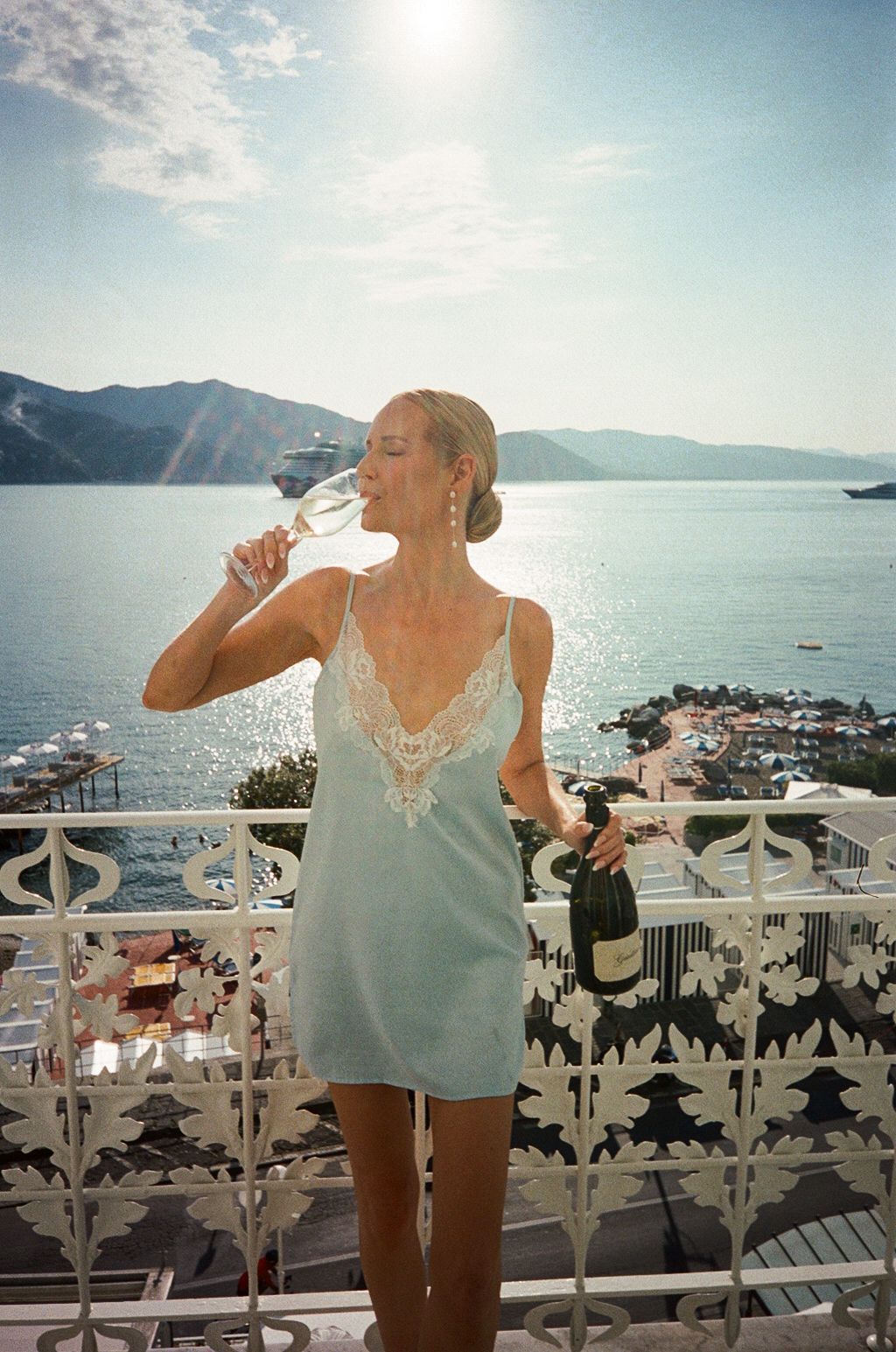 Woman in blue slip dress sipping champagne on a scenic balcony overlooking the Mediterranean sea.