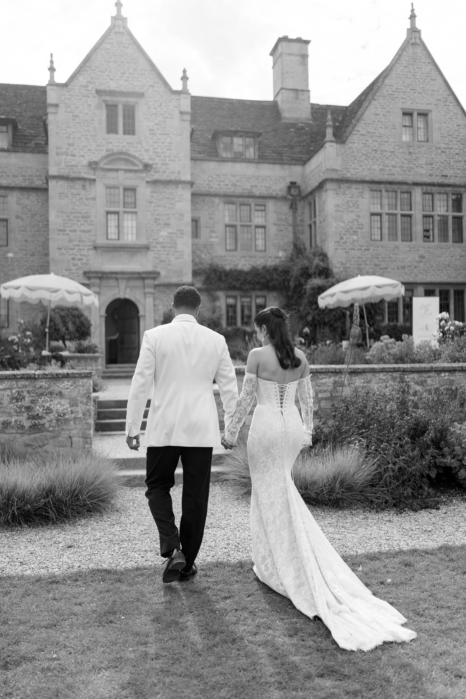 Bride in fitted lace gown with train and groom in white jacket walking toward a stone manor house.