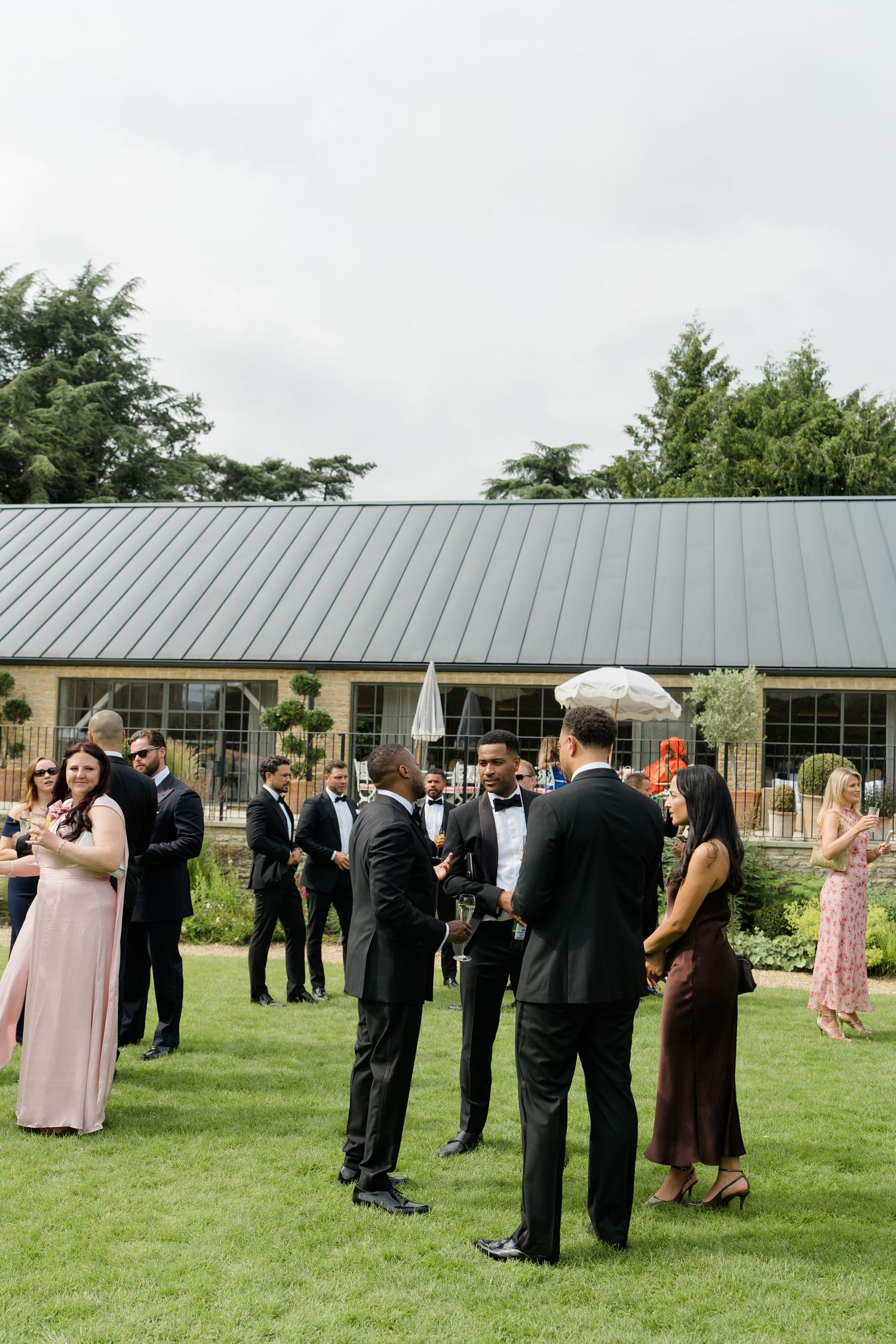 Wedding guests mingling on manicured lawn outside modern venue with metal roof on overcast day.