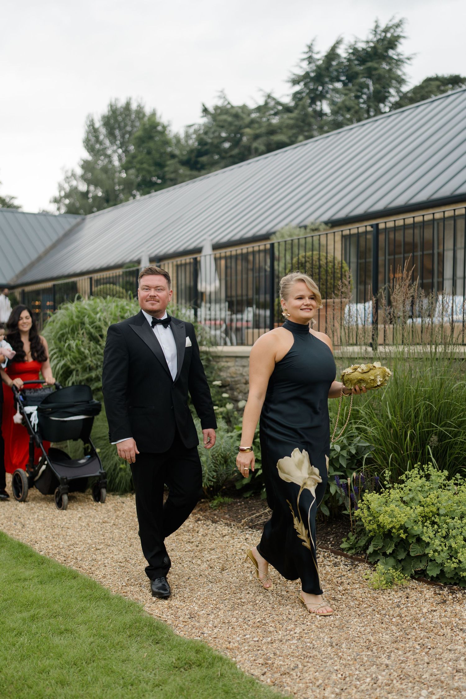 Elegant couple in black tie attire walking along garden path at wedding venue.