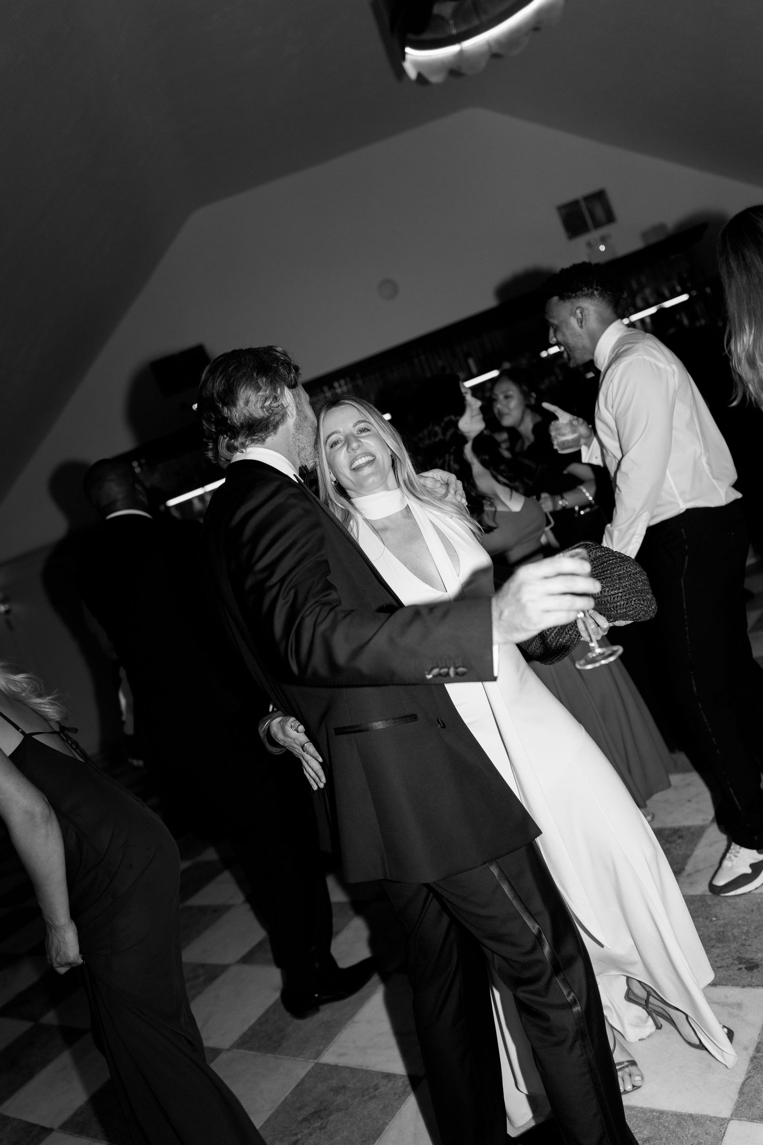 Black and white photo of a laughing bride holding wine glass while dancing with groom at reception.