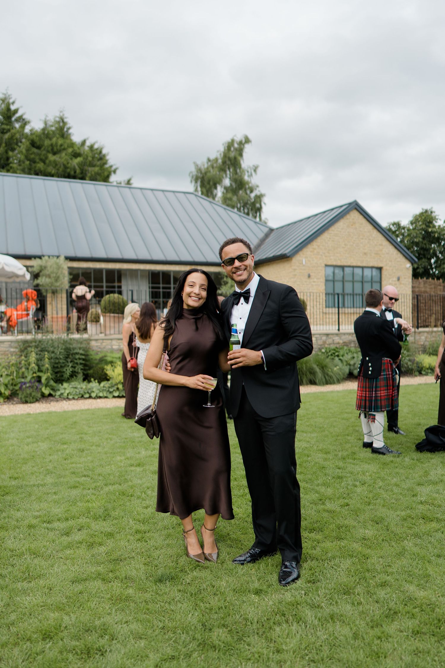 A couple in formal attire smile together on a lawn with a bagpiper visible in the background.
