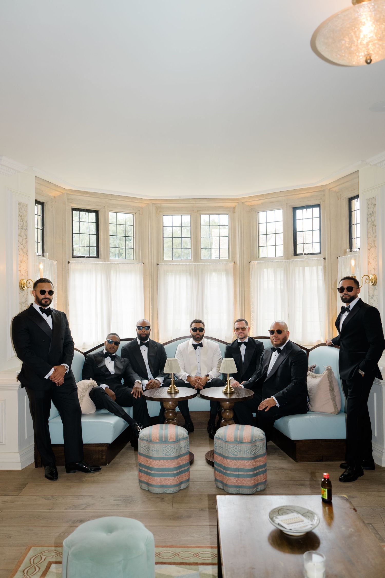 Six groomsmen in black tuxedos posing in elegant bay window sitting room.