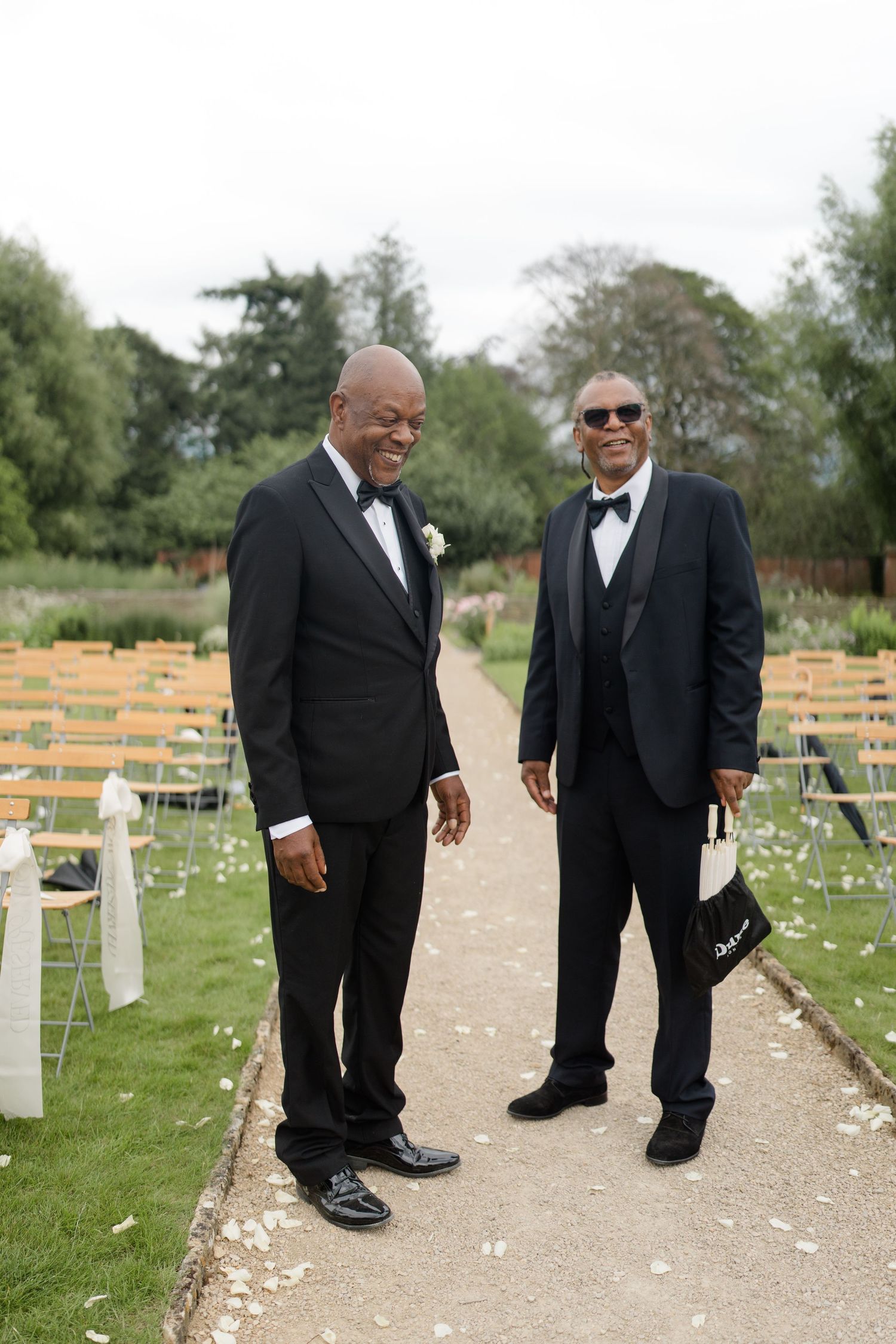 Two elegantly dressed men in black tuxedos smile at an outdoor wedding ceremony venue.