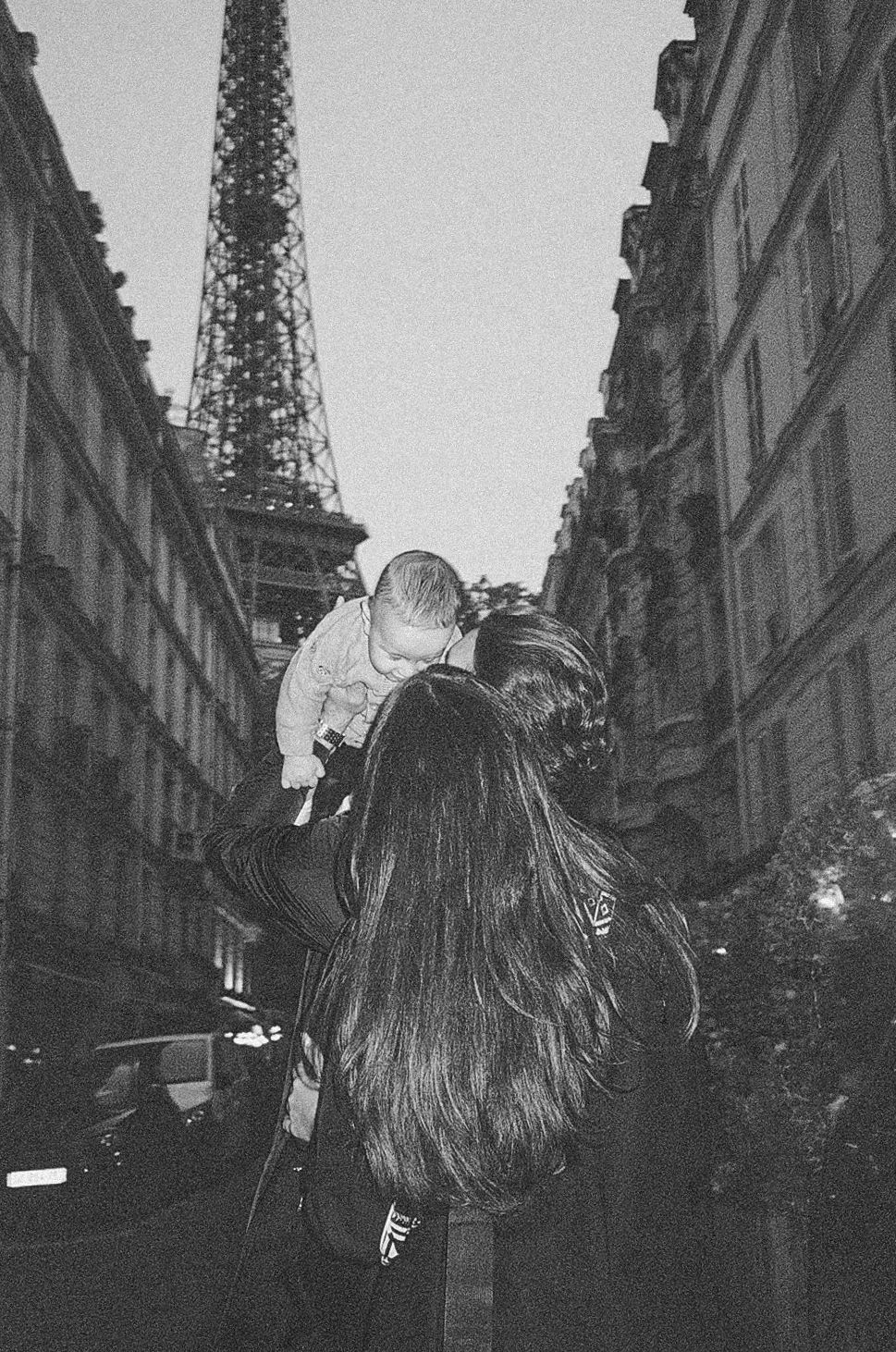 Black and white photo of a woman lifting a baby near the Eiffel Tower on a Parisian street.