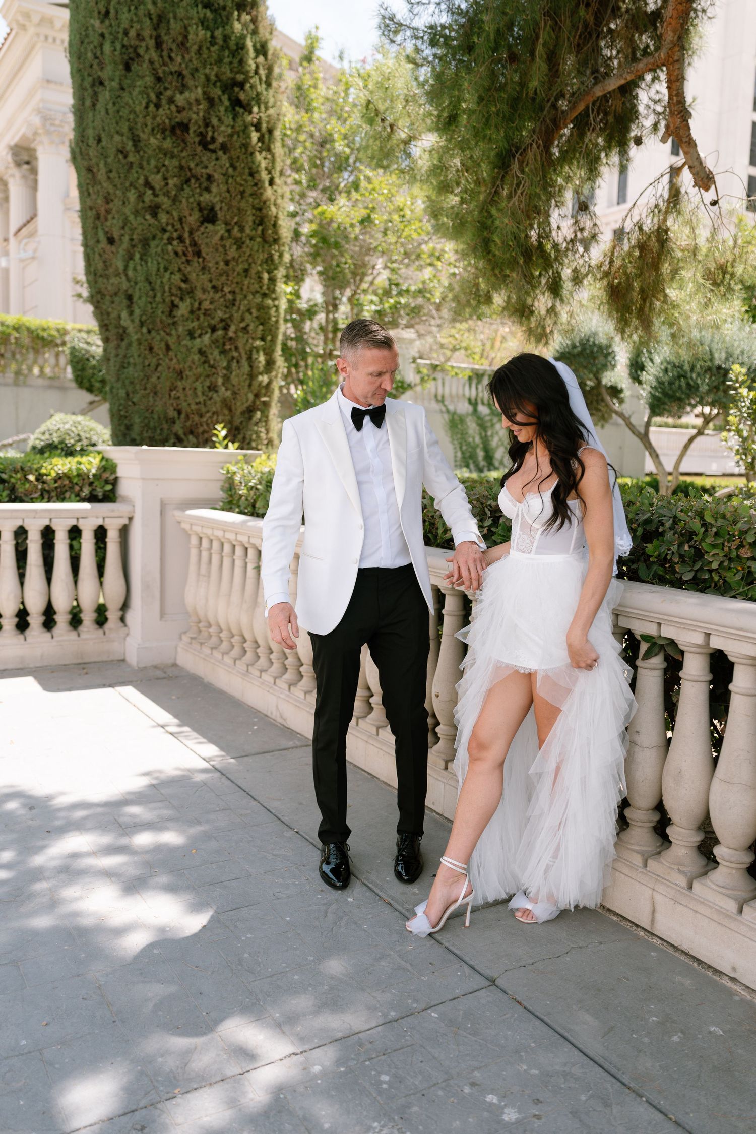A wedding couple poses on an elegant stone terrace with cypress trees.