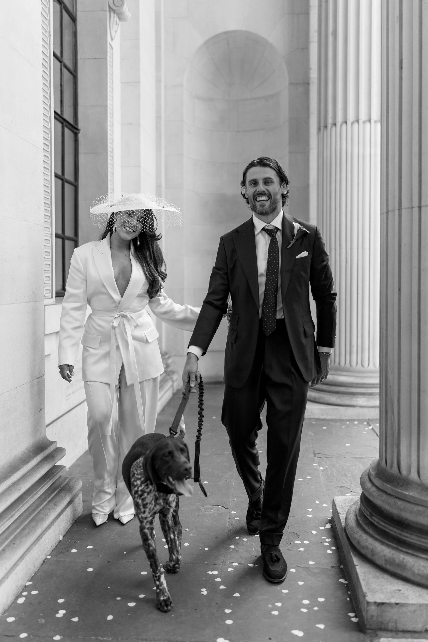 Bride in white suit and hat walks with groom and spotted dog between grand columns.