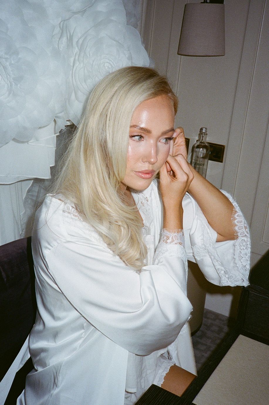 Blonde woman in white satin robe with lace trim poses thoughtfully in elegant room with soft lighting.