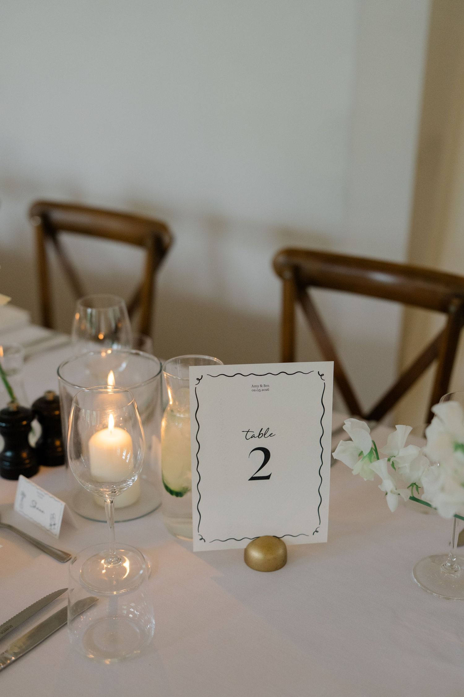 Elegant wedding table number two card beside lit candle and white floral arrangement on white tablecloth.