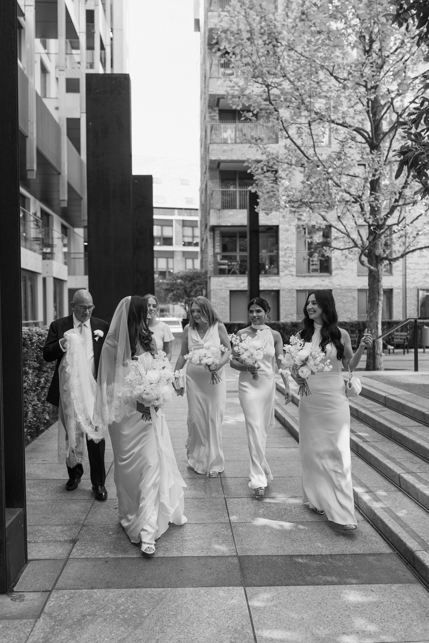 Bride in veil walks with bridesmaids in satin gowns holding bouquets through urban courtyard in black and white.