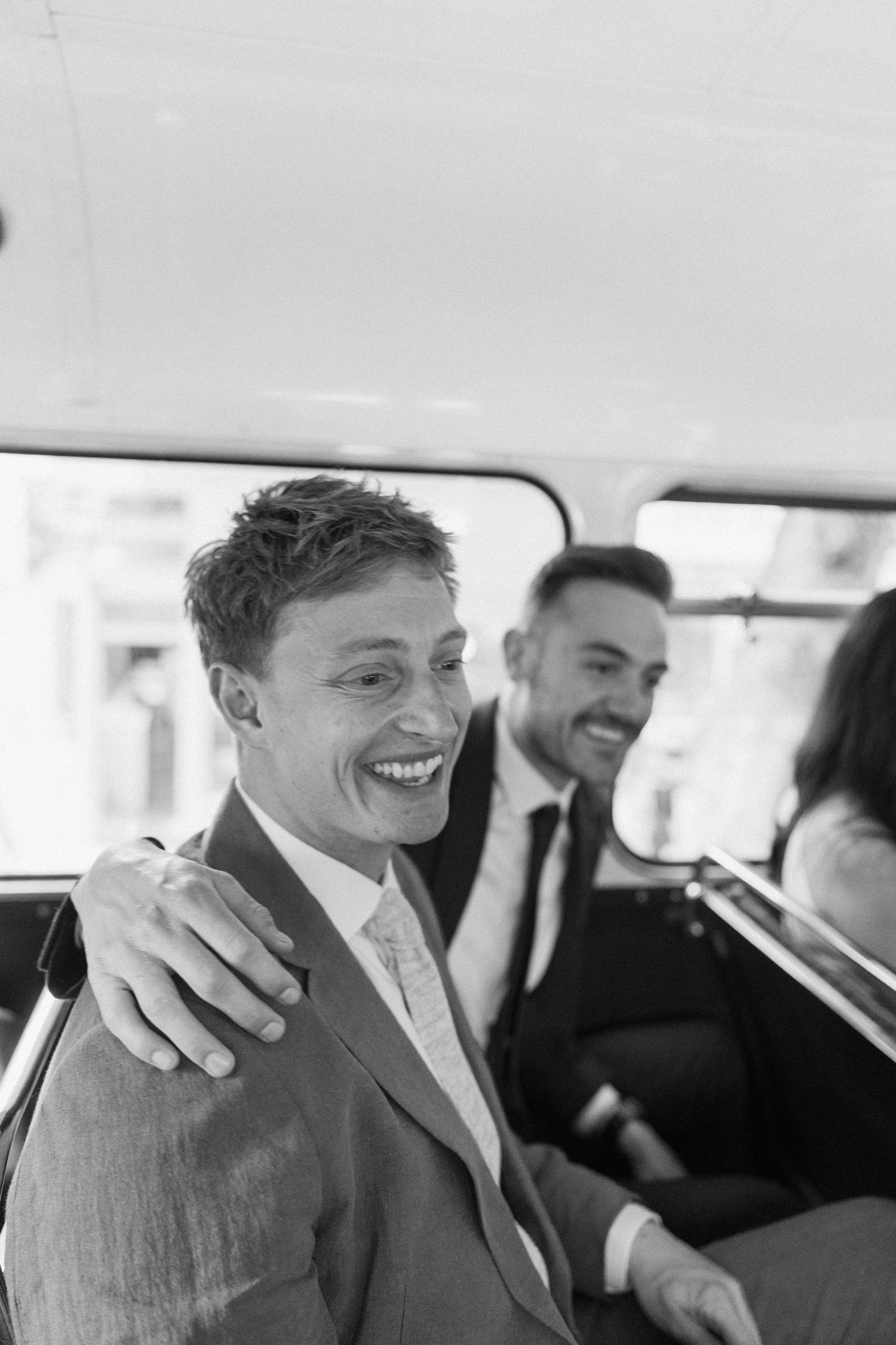 Black and white photo of a smiling groom laughing with groomsmen inside a vintage vehicle on his wedding day.