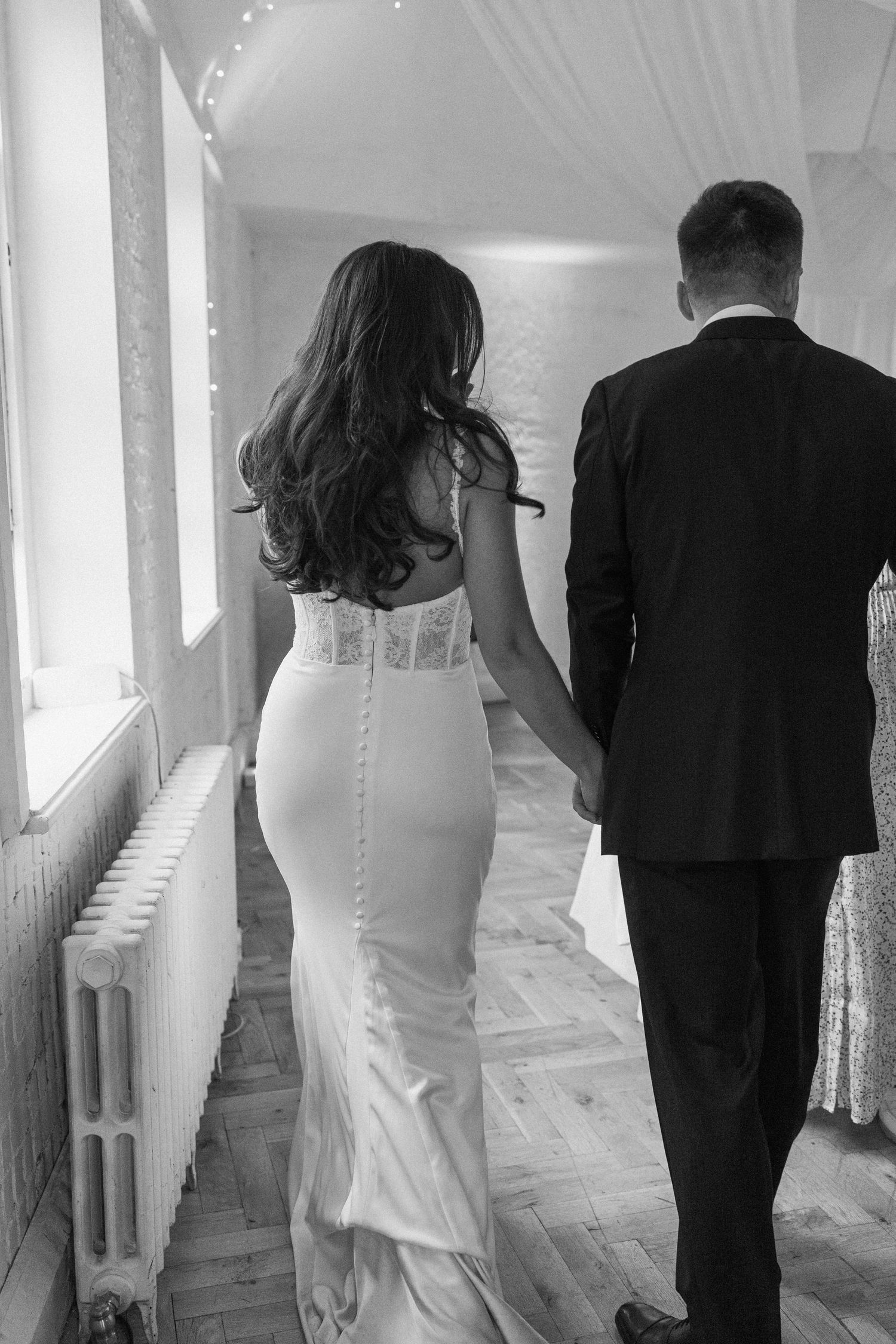 Black and white rear view of bride in fitted lace gown holding hands with groom walking down a hallway.