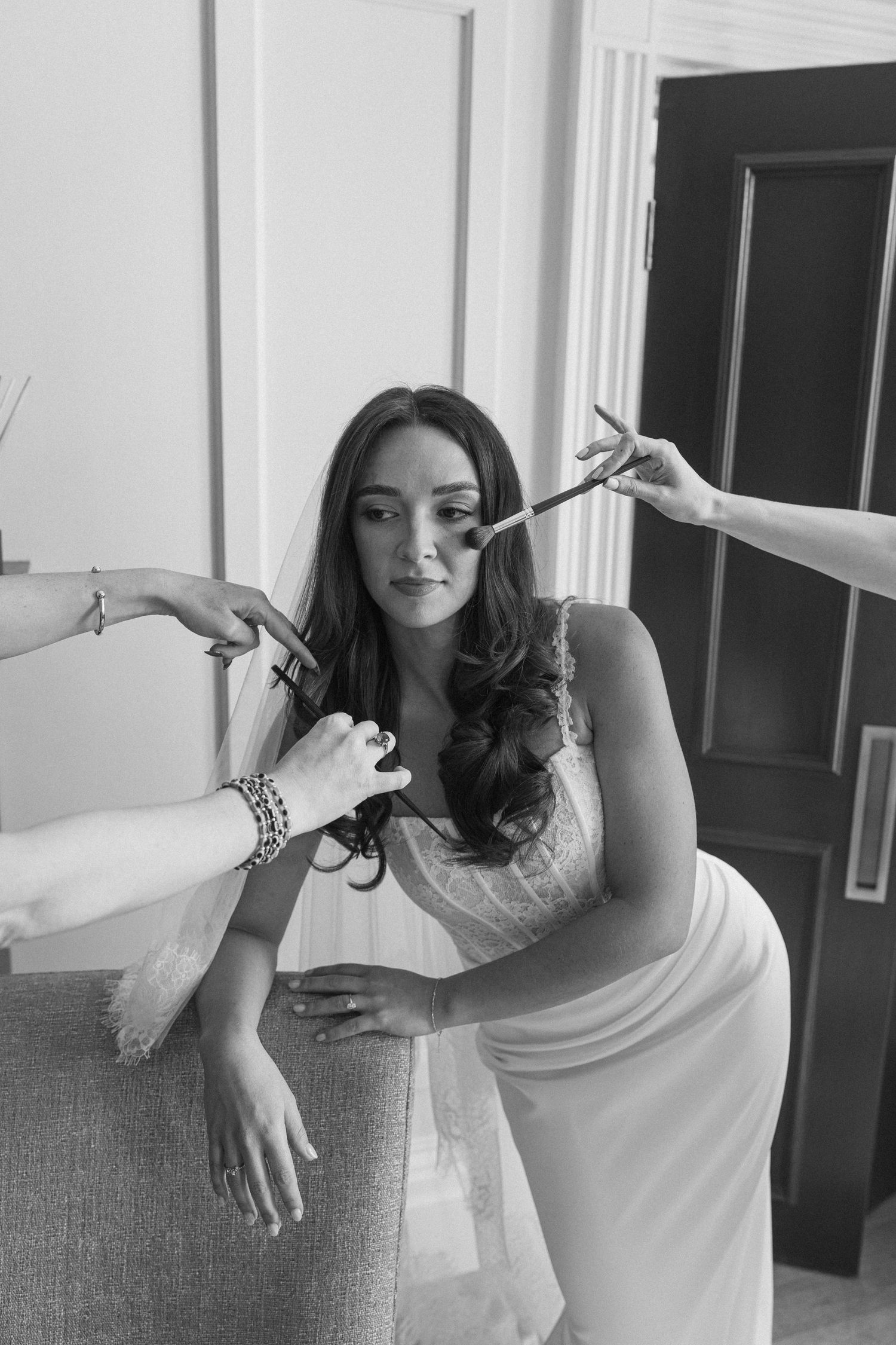Black and white photo of a bride in a white gown being pampered by makeup and hair stylists.