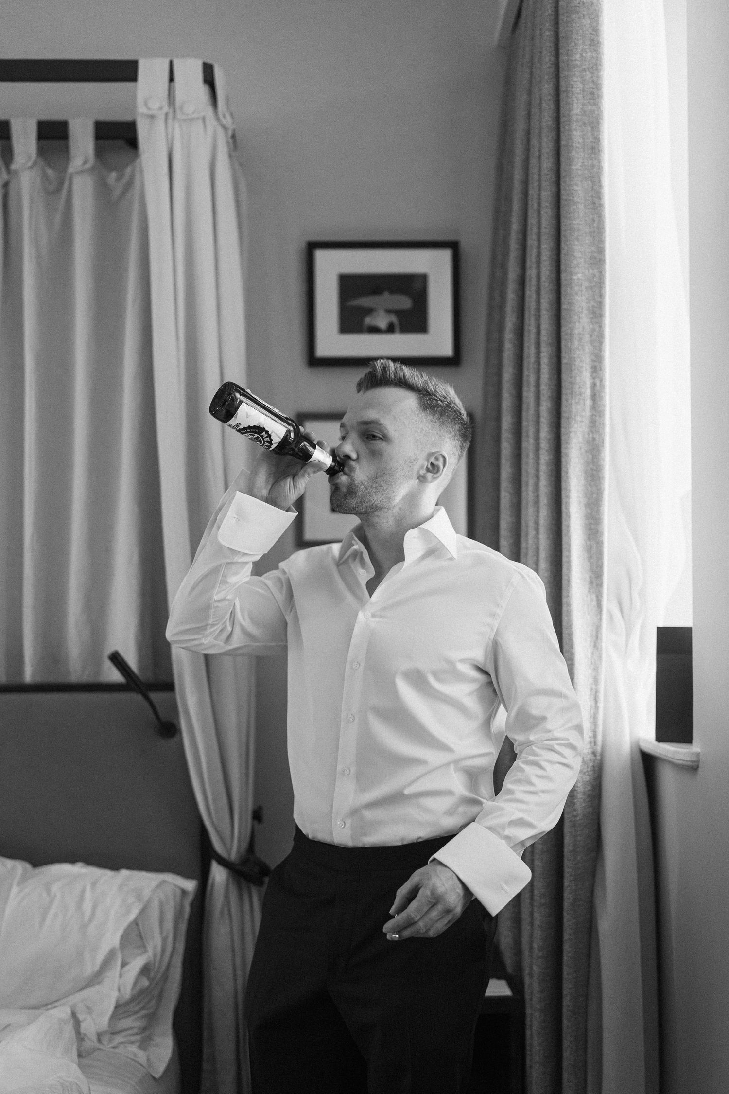 Man in white dress shirt drinking from a bottle in a hotel room, black and white photography.