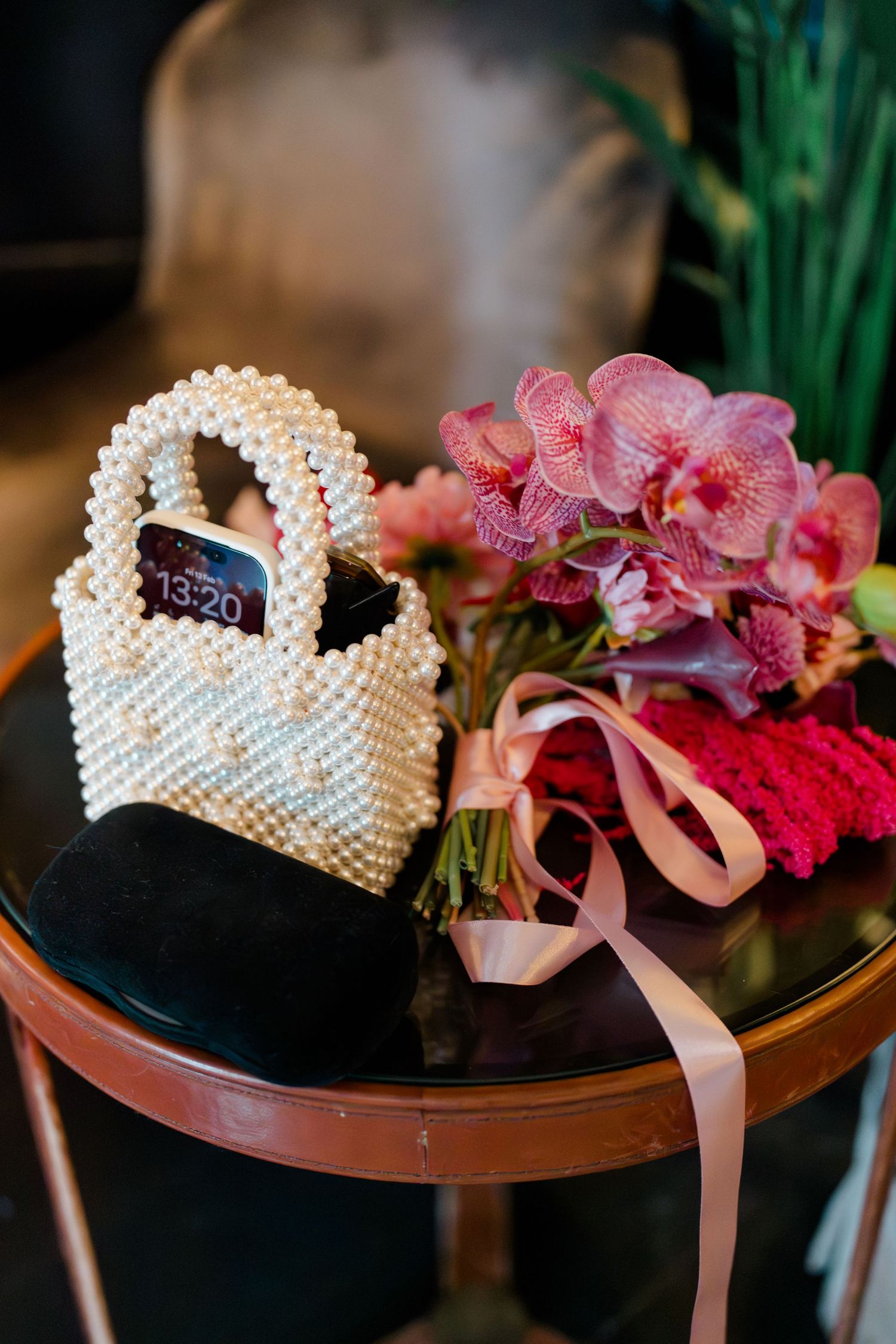 Pearl beaded handbag with phone beside pink orchid wedding bouquet with satin ribbon on table.