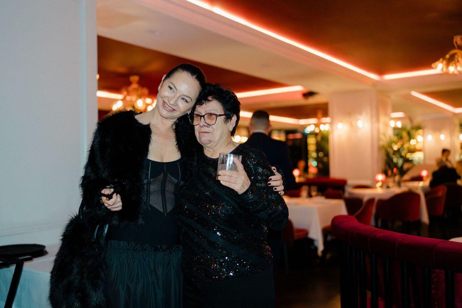 Two women in black outfits smiling together at an elegant warmly lit wedding reception venue.