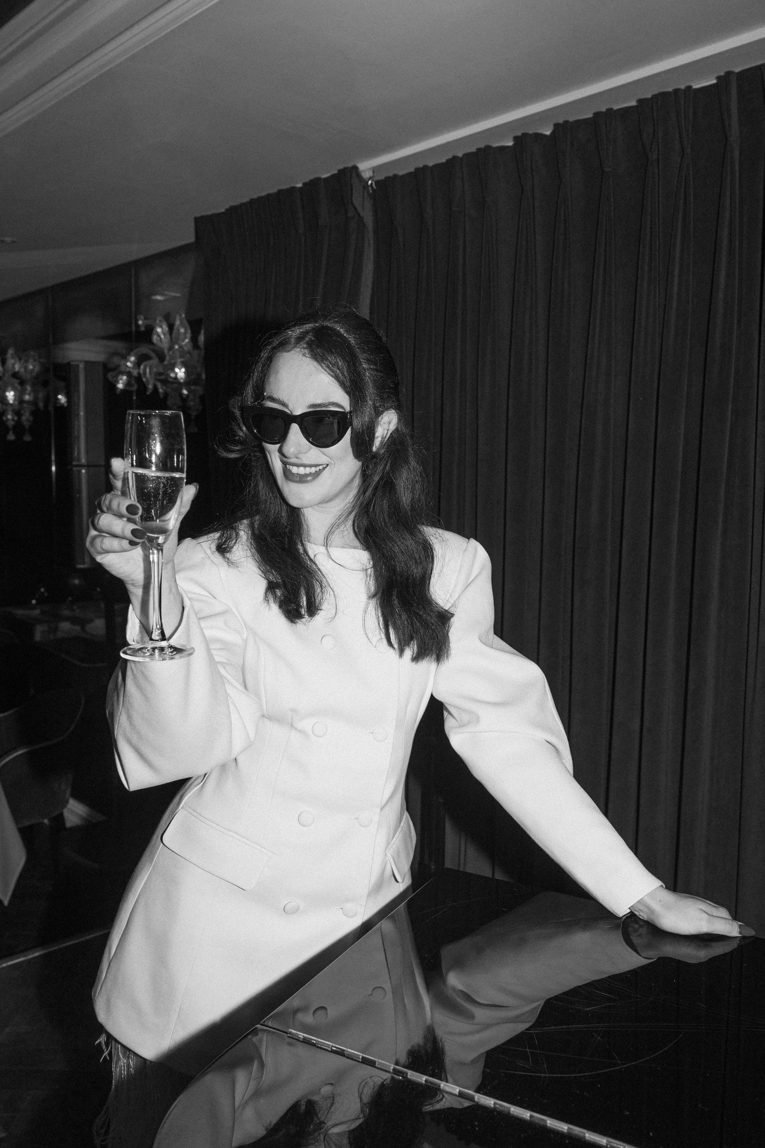 Black and white photo of a radiant bride in white blazer raising a champagne glass in celebration.