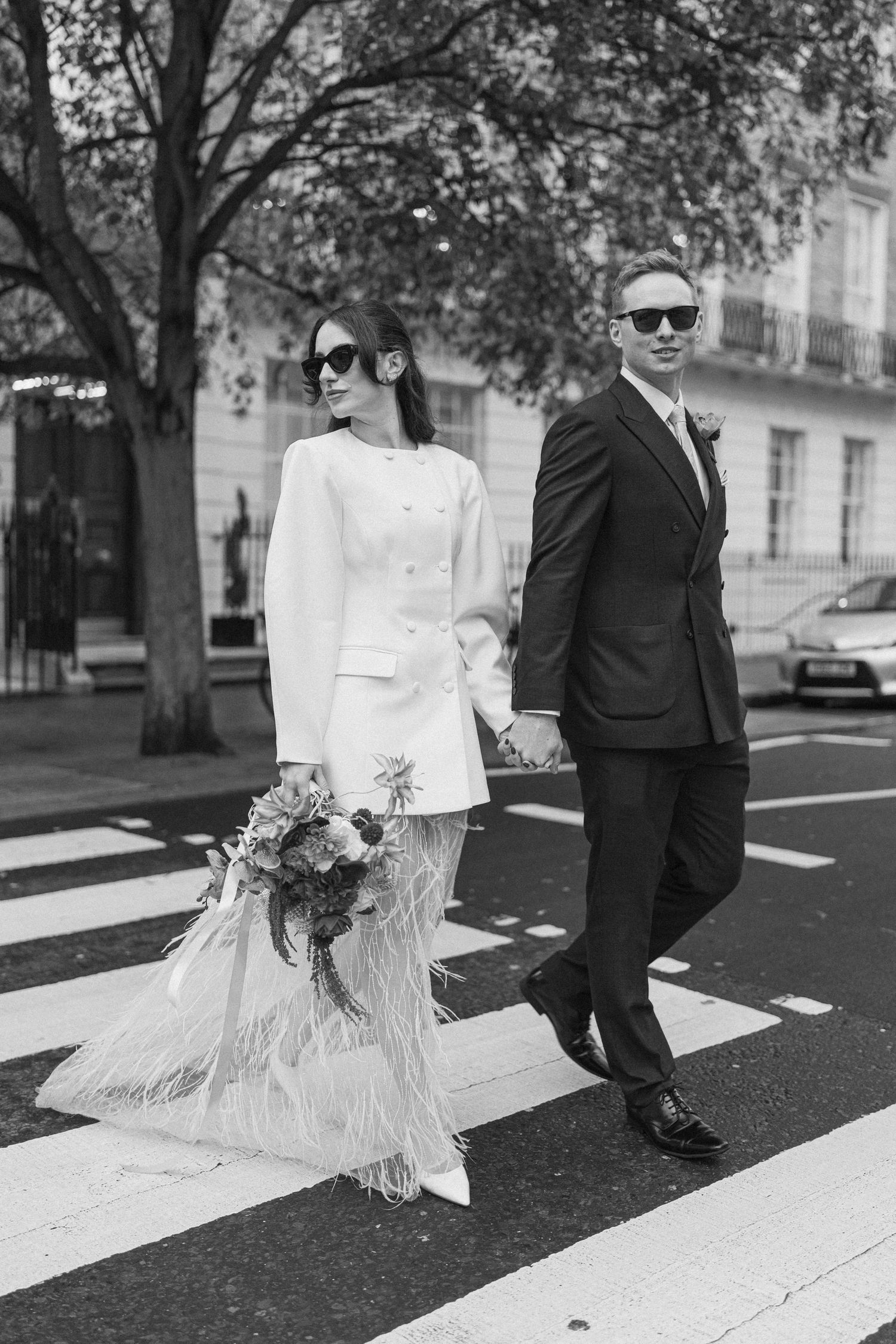 Black and white photo of stylish bride and groom in sunglasses crossing a zebra crossing hand in hand.