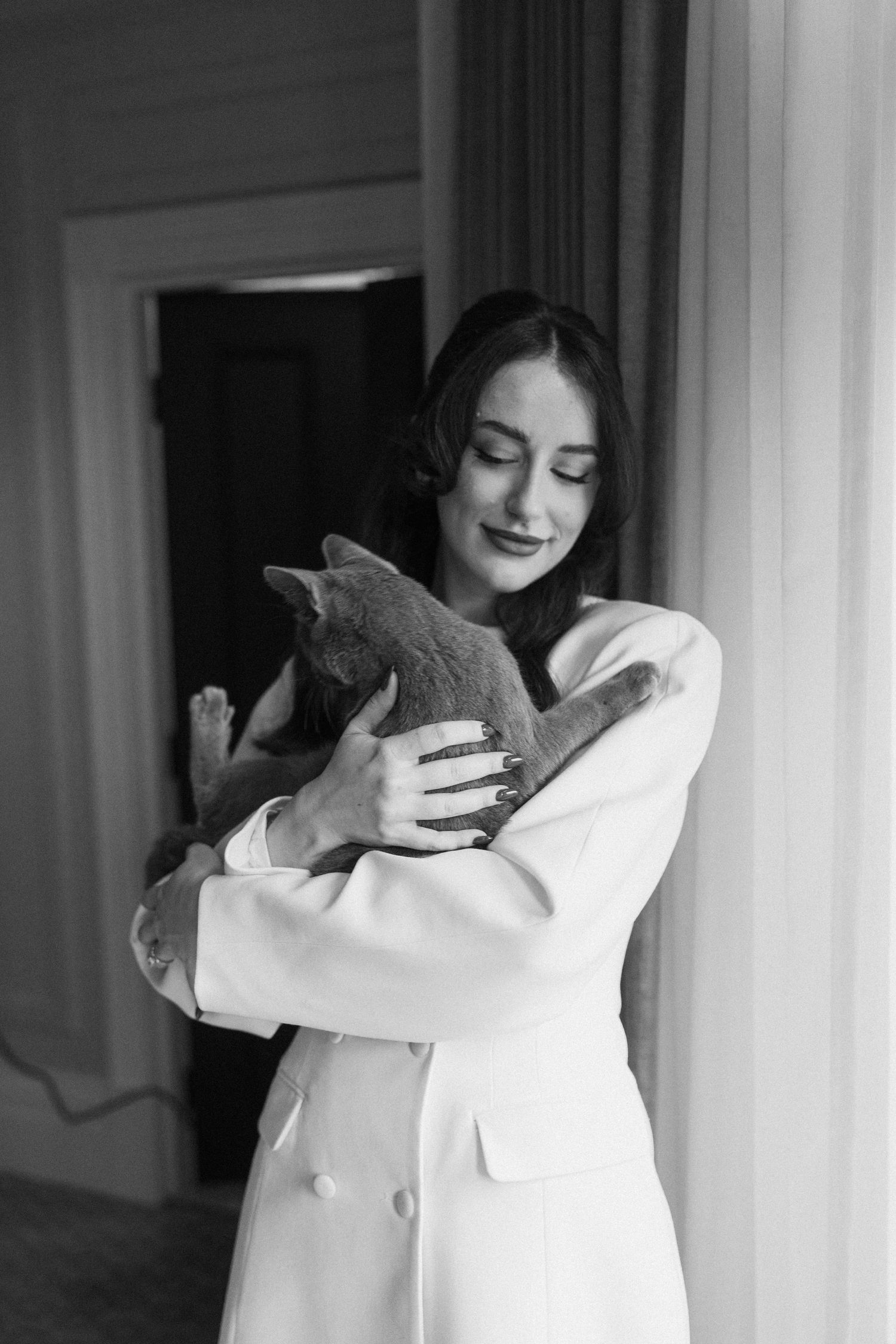 Black and white photo of bride in white blazer dress tenderly holding a gray cat.