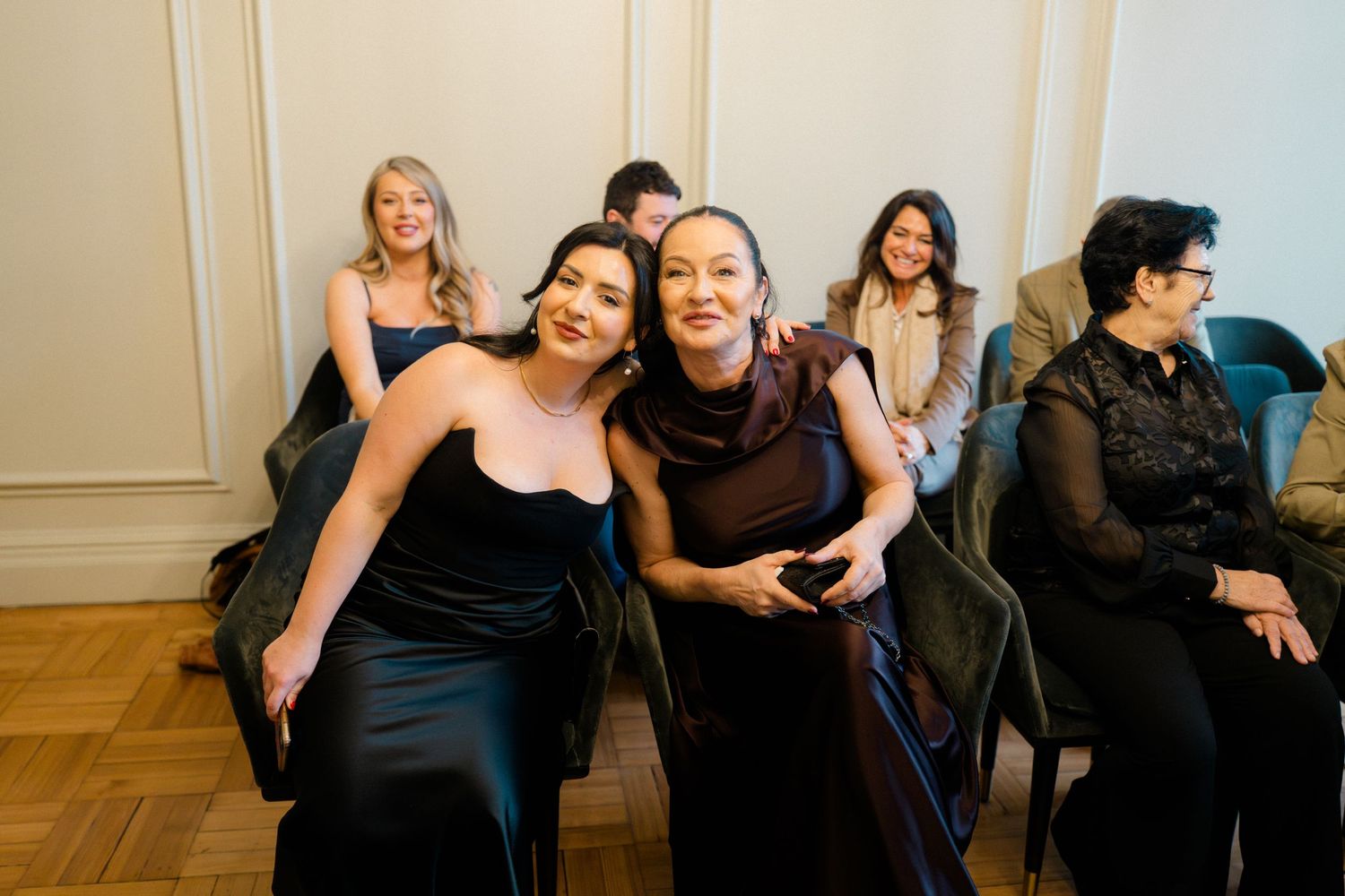 Group of smiling women in dark formal dresses seated at an intimate indoor wedding ceremony.