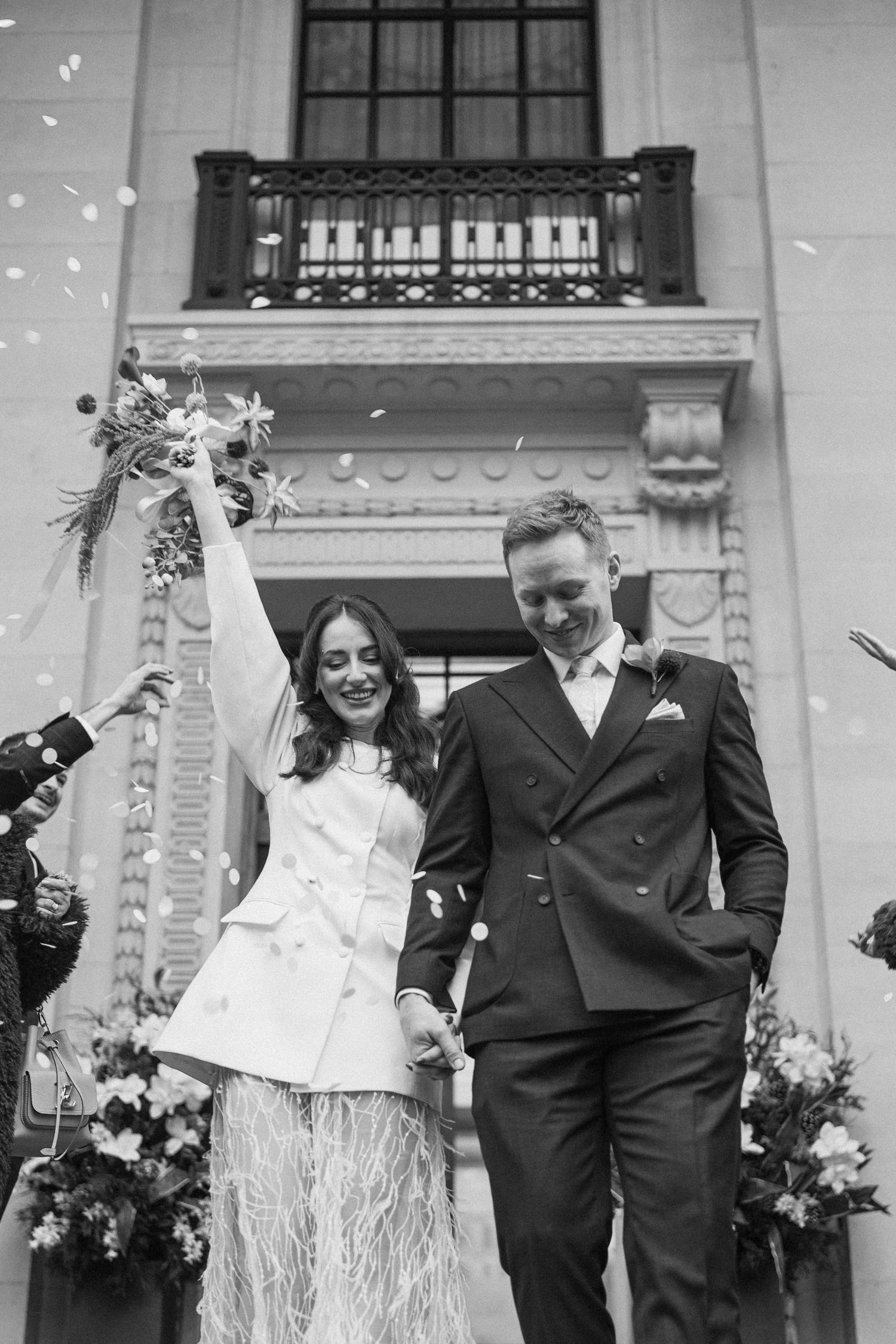 Black and white photo of newlyweds walking out of ceremony venue in confetti shower.