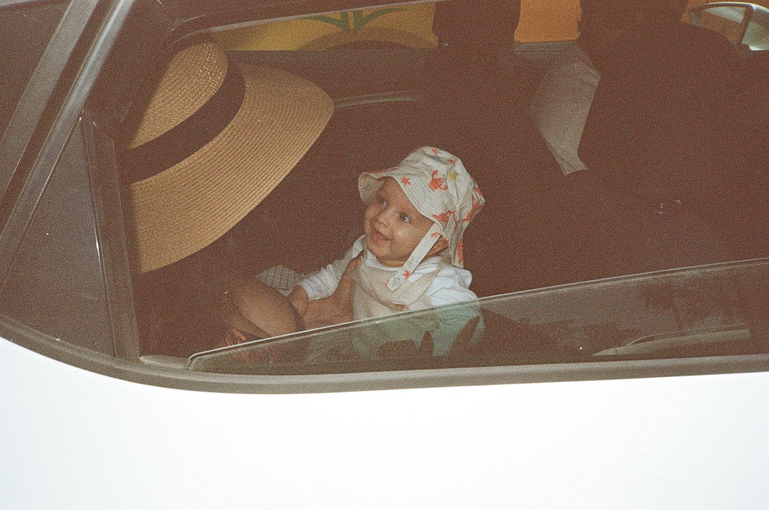 Baby wearing a floral hat sitting in a car seat inside a vehicle, with a large straw hat visible in the background.