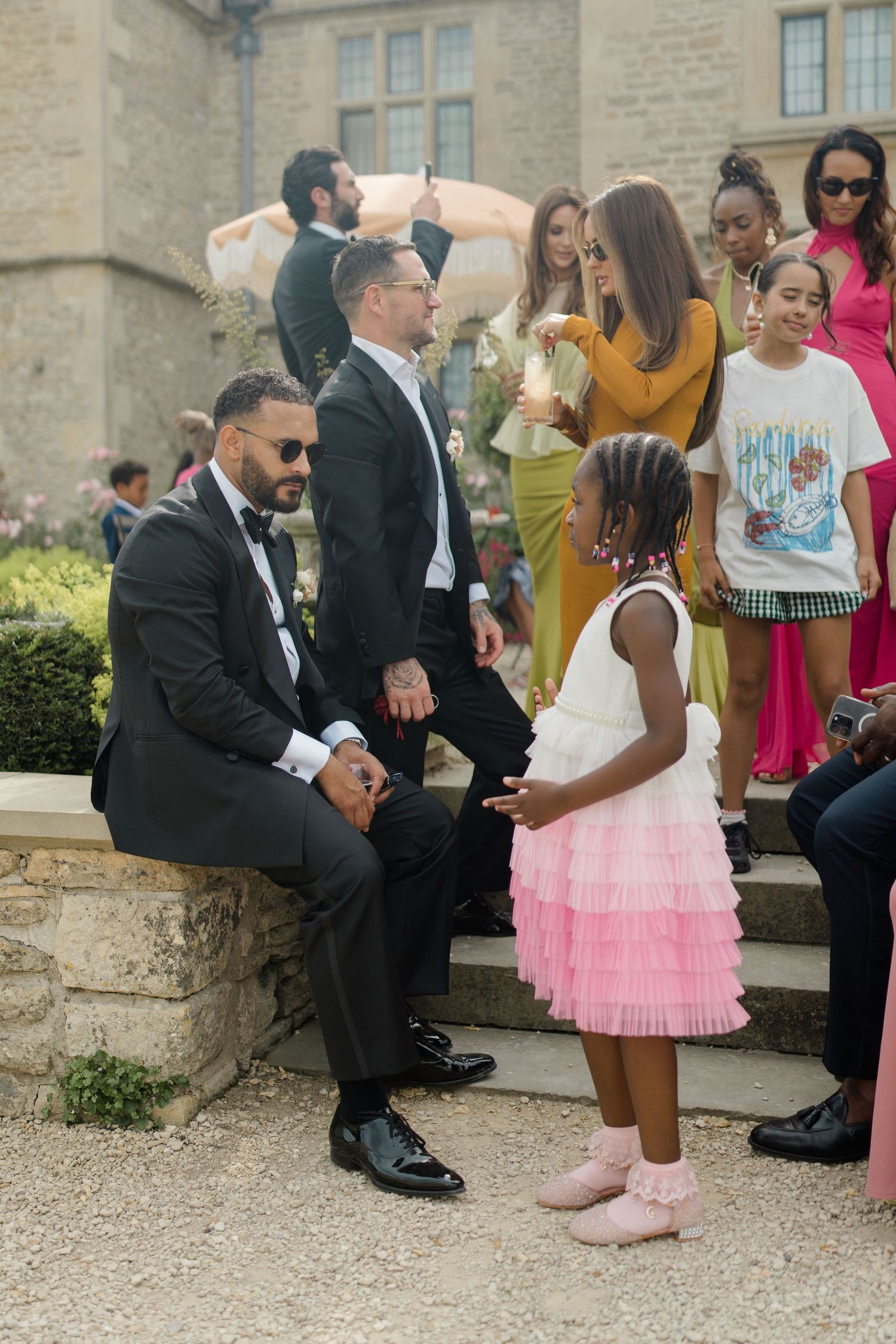 Groom in tuxedo chats with young girl in pink dress on stone steps at outdoor wedding reception.
