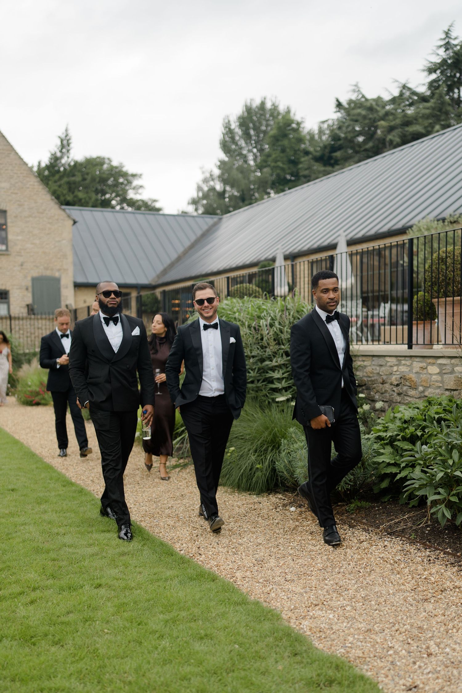 Groomsmen in black tuxedos with bow ties walking confidently along garden path at wedding venue.