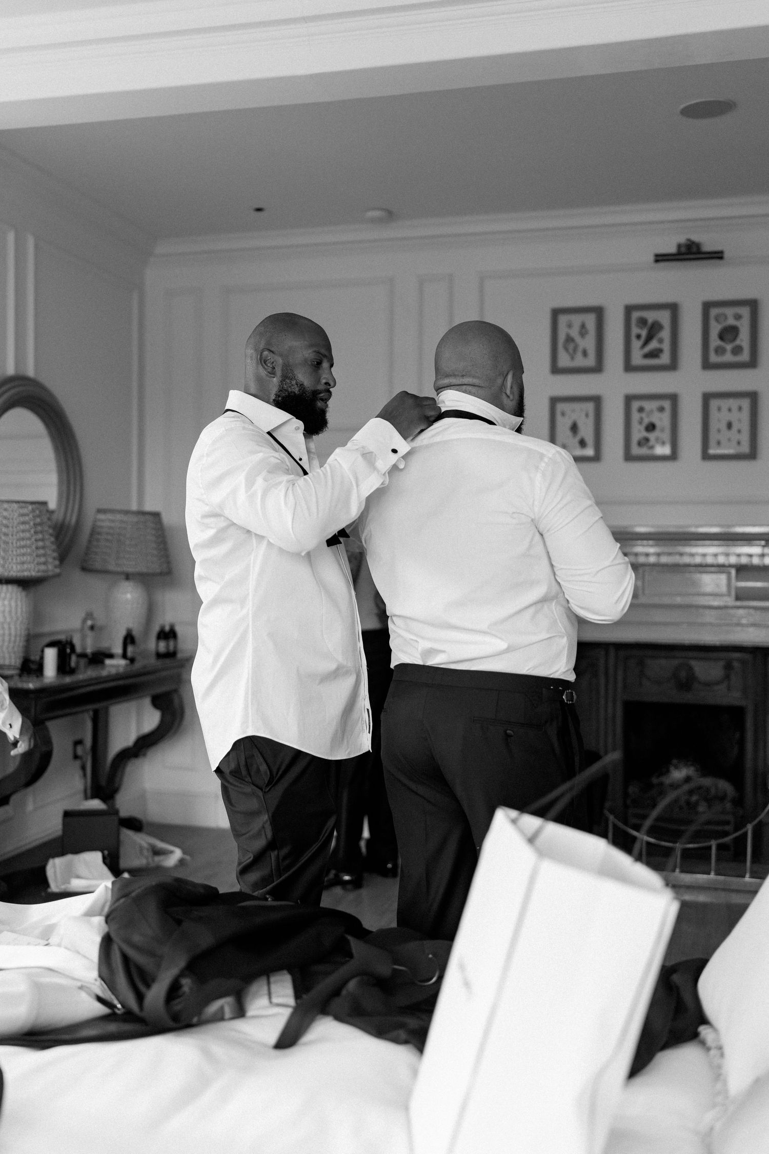 Two groomsmen in white dress shirts help each other with bow ties while getting ready for a wedding.