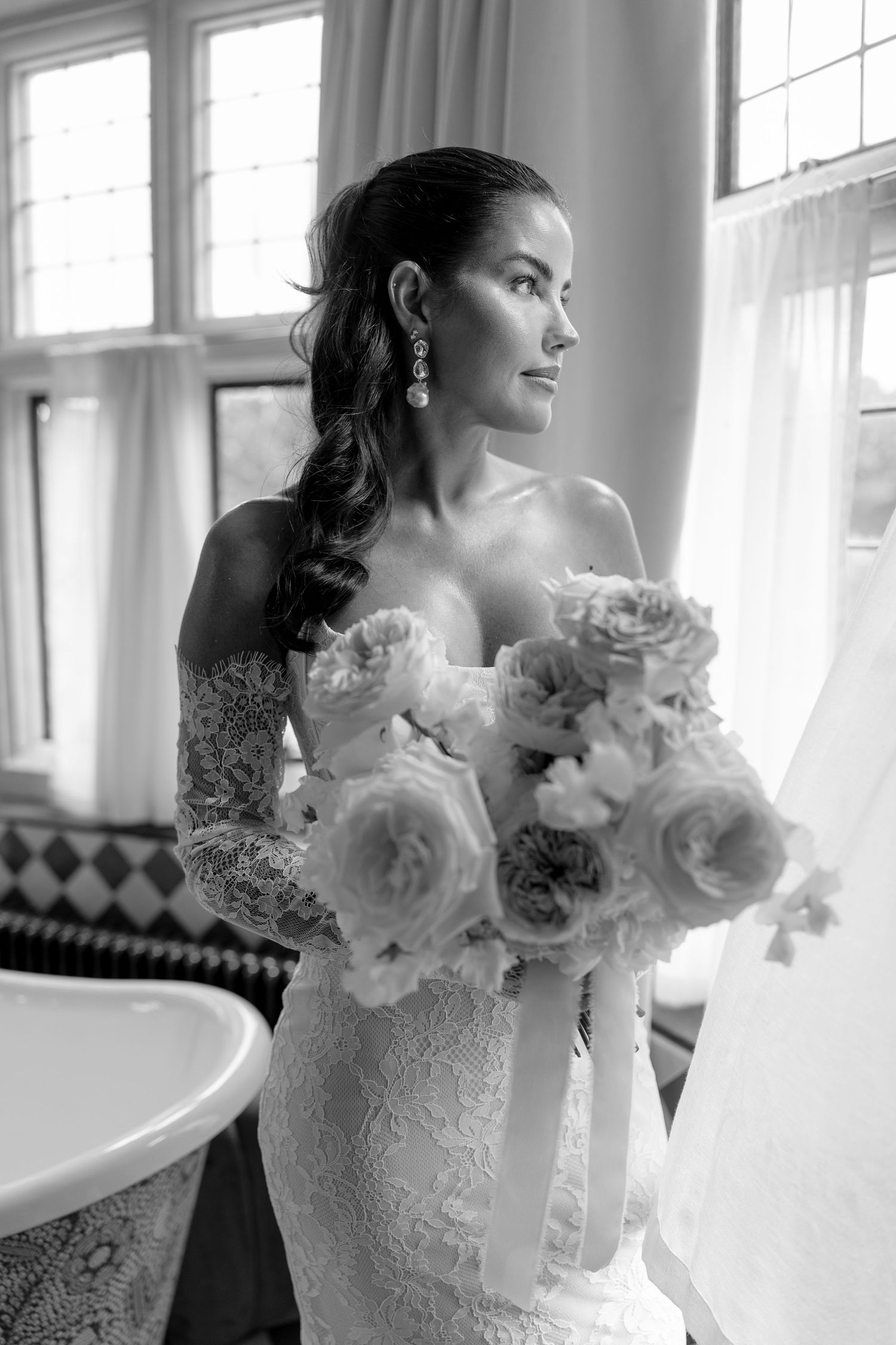 Beautiful bride in lace off-shoulder gown holds peony bouquet by window in black and white photo.