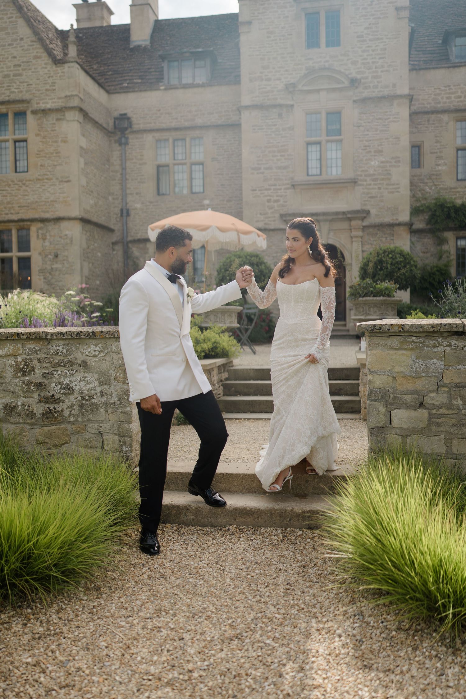 Bride and groom walk hand in hand through garden path of historic stone manor estate.